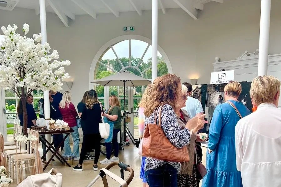 People shopping at a market or boutique inside a bright, airy space with large arched windows, white walls, and decorative white columns, surrounded by floral decor and display tables.