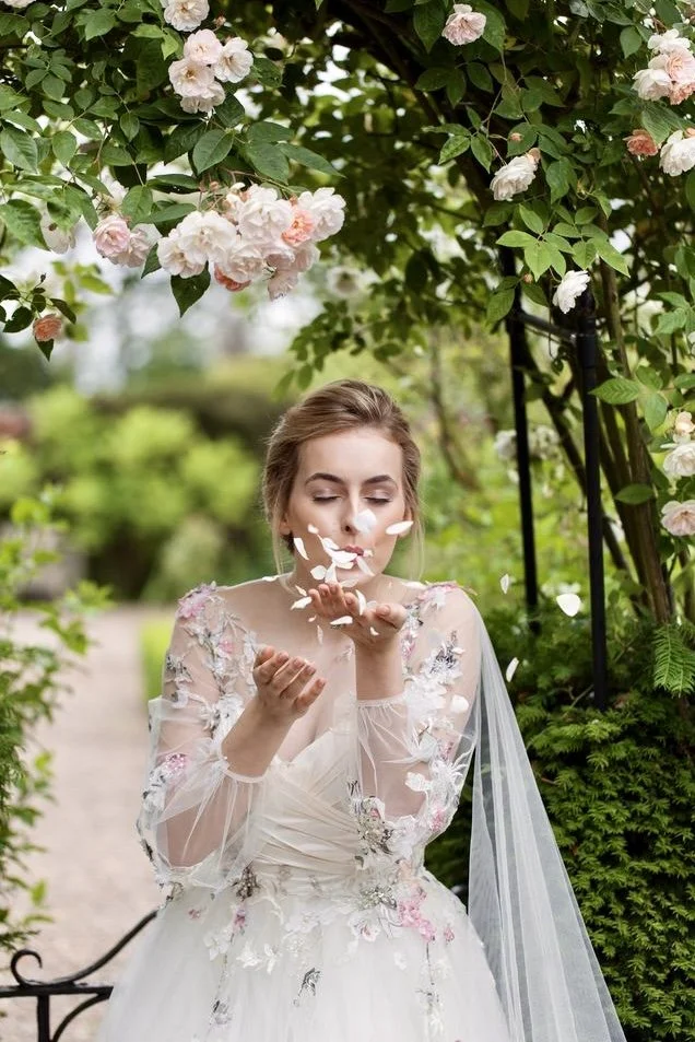 A woman in a wedding dress blows flower petals in a garden with pink and white flowers overhead.