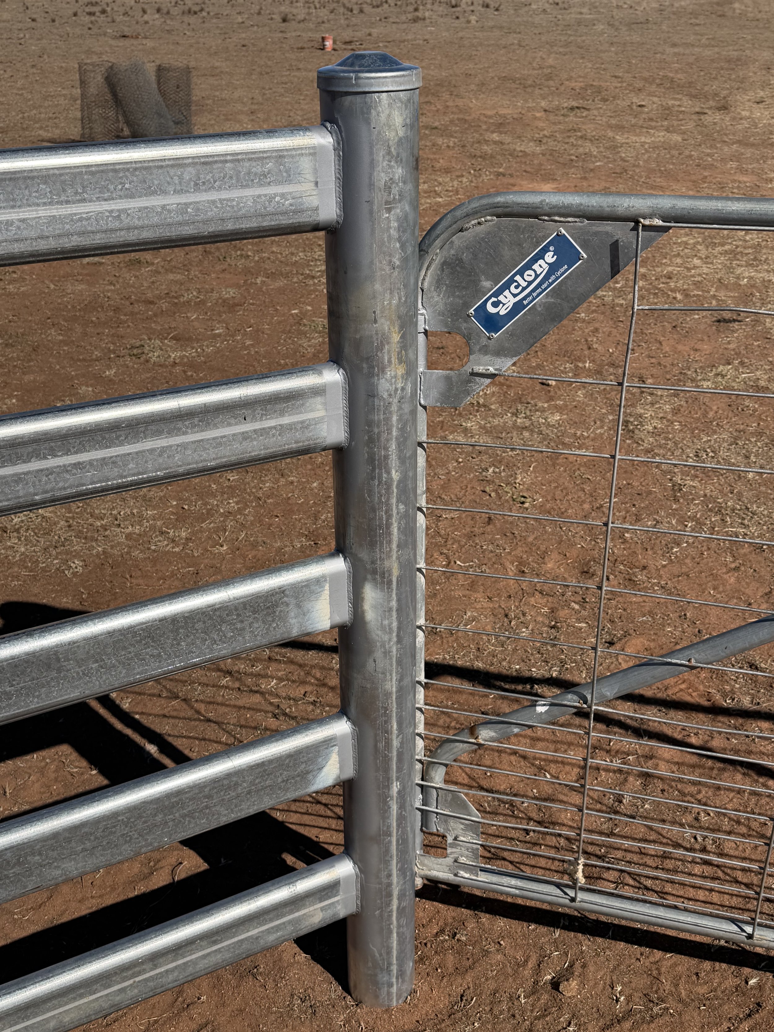 Close-up of a metal livestock gate with a blue Cyclone sign, set on dirt ground with dry grass and small objects in the background.