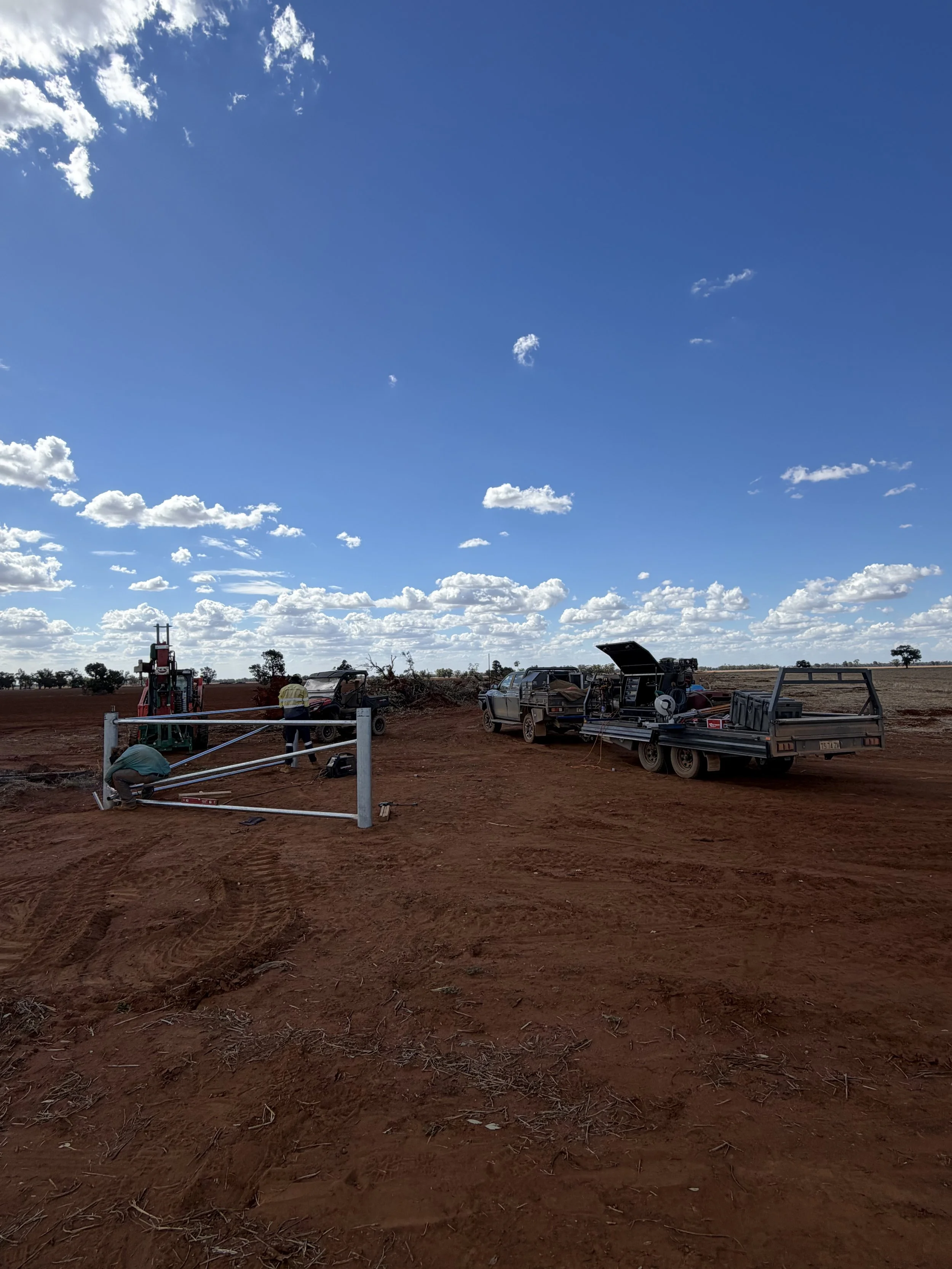 Workers setting up a metal fence on a red dirt field with vehicles and equipment nearby, under a blue sky with scattered white clouds.