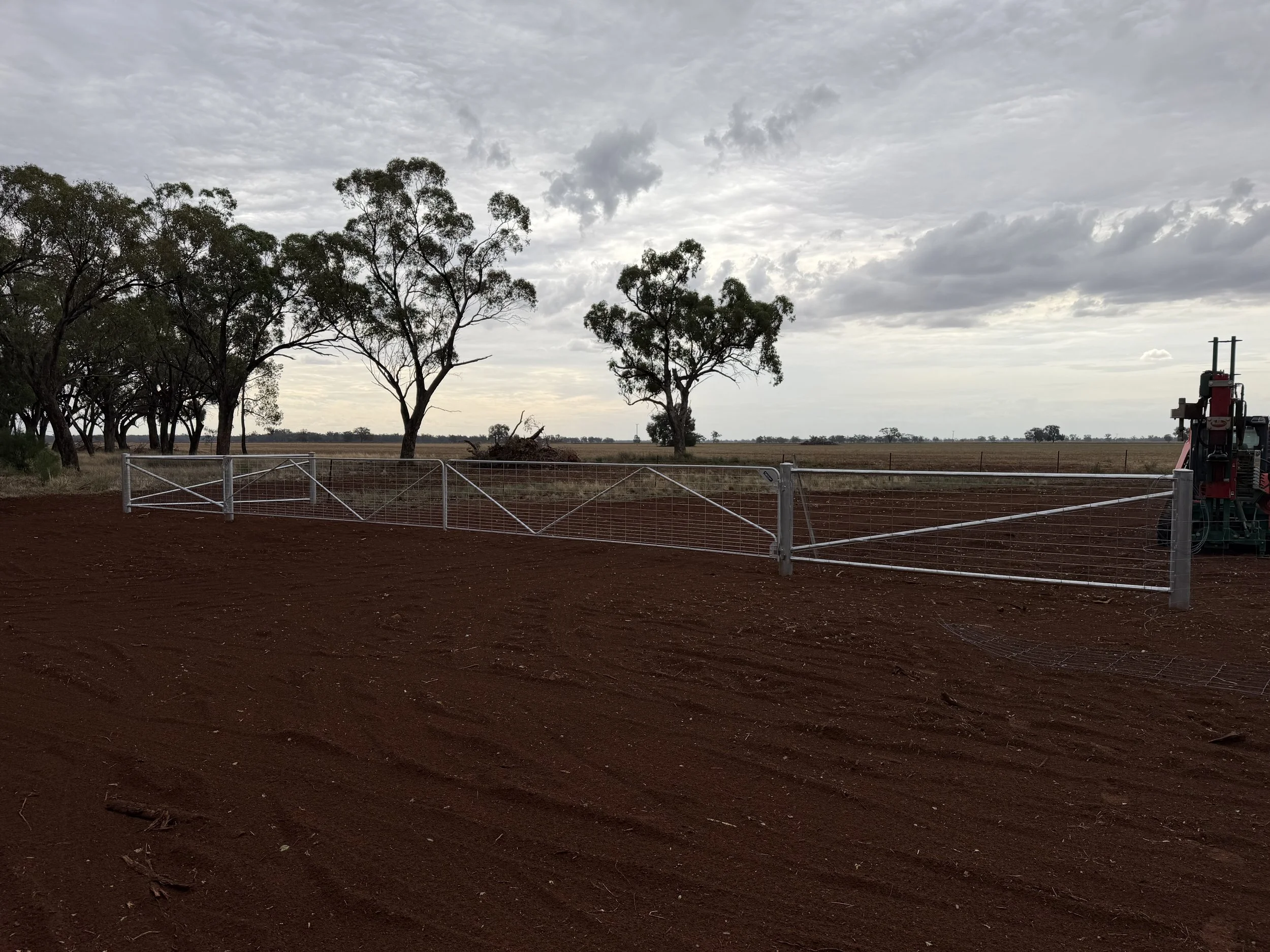 A rural landscape with several large trees, a cloudy sky, and a metal fence in the foreground. There is some farm machinery partially visible on the right side of the image.