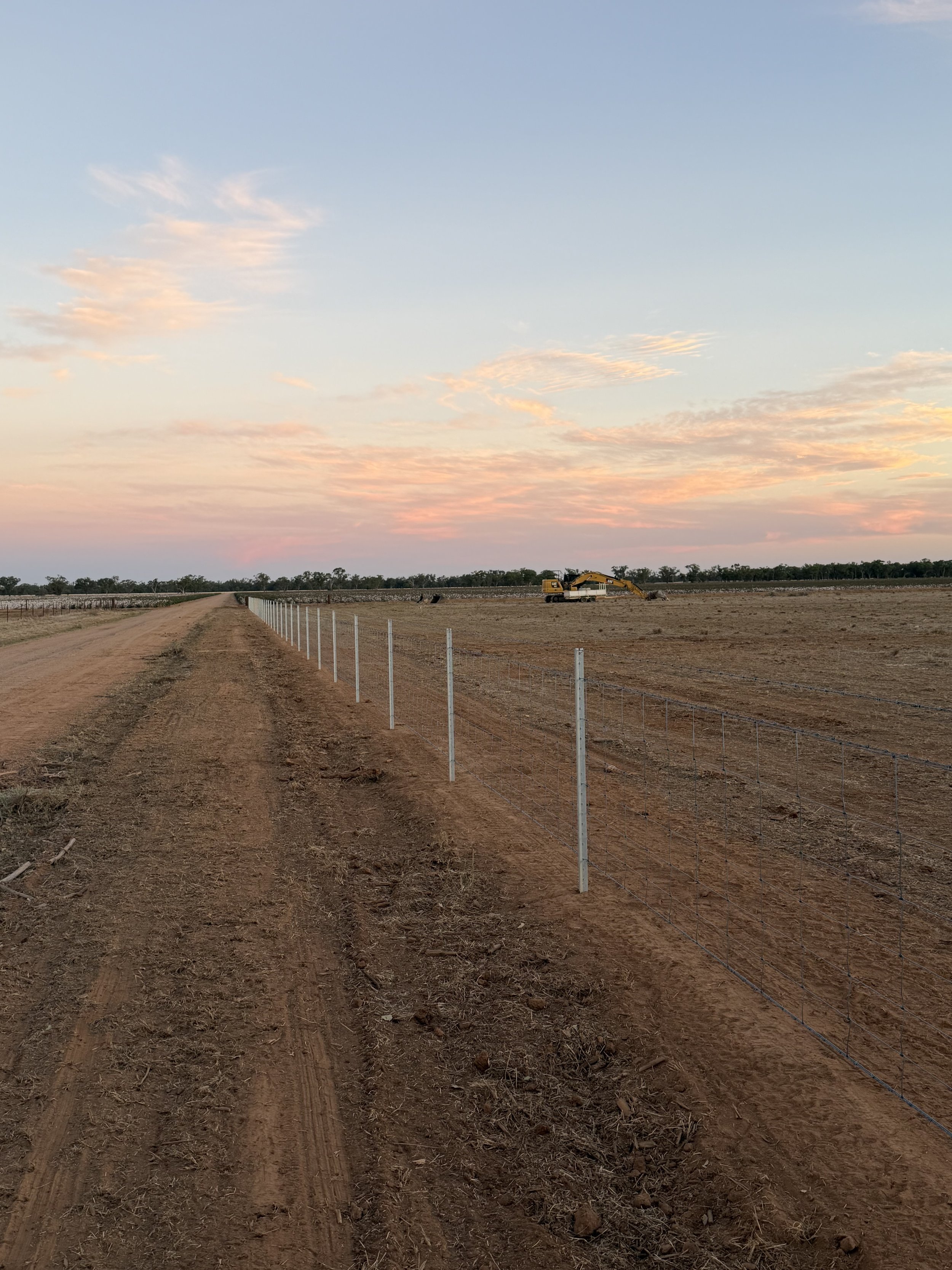 A construction site with a dirt road, a yellow excavator, and a fence, under a sky with pink and blue clouds at sunset.