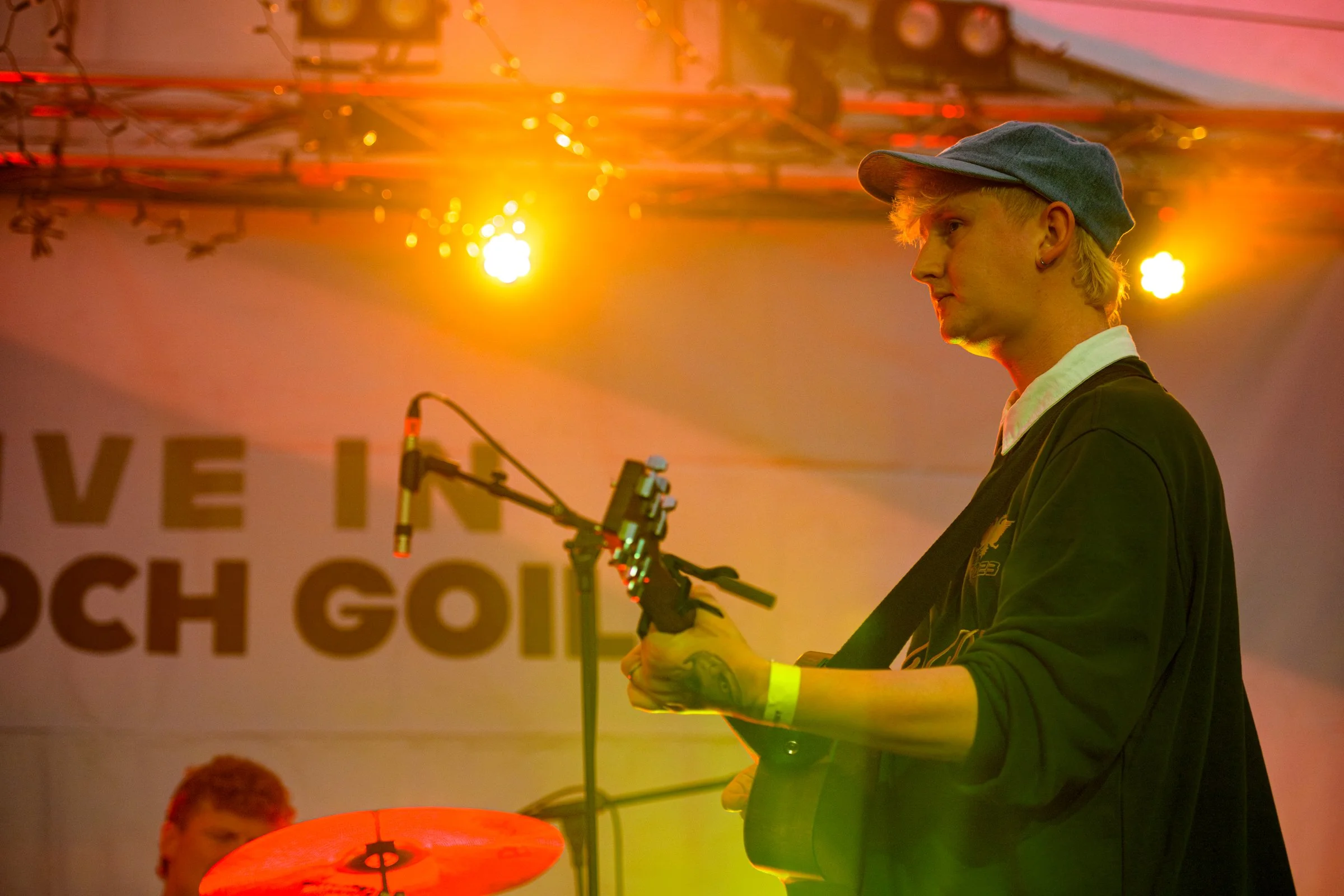 A person with blonde hair wearing a cap and earring, holding a microphone stand, is on stage with orange and yellow lighting. In the background, a sign with partially visible text is displayed.