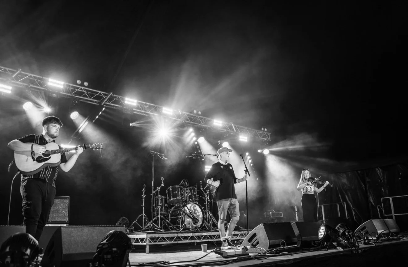 Black and white photo of a band performing live on stage at night, with bright stage lights overhead and musical instruments including a drum set, a guitar, and a violin.