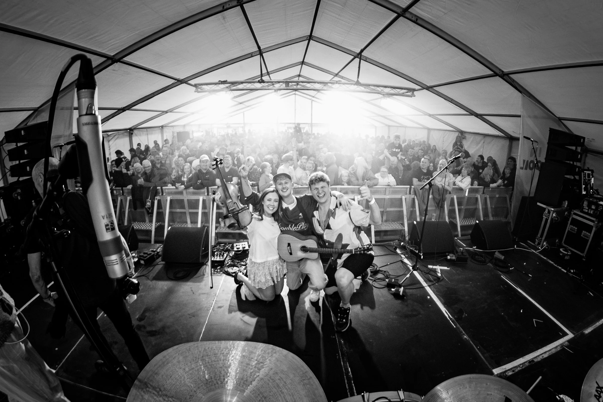 A group of people on a stage holding musical instruments, with a large audience in the background under a tent. The image is in black and white.