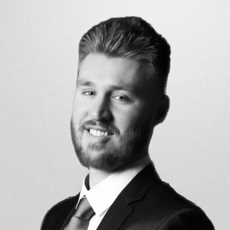 Black and white professional portrait of a young man with styled hair and a beard, wearing a suit and tie, smiling.