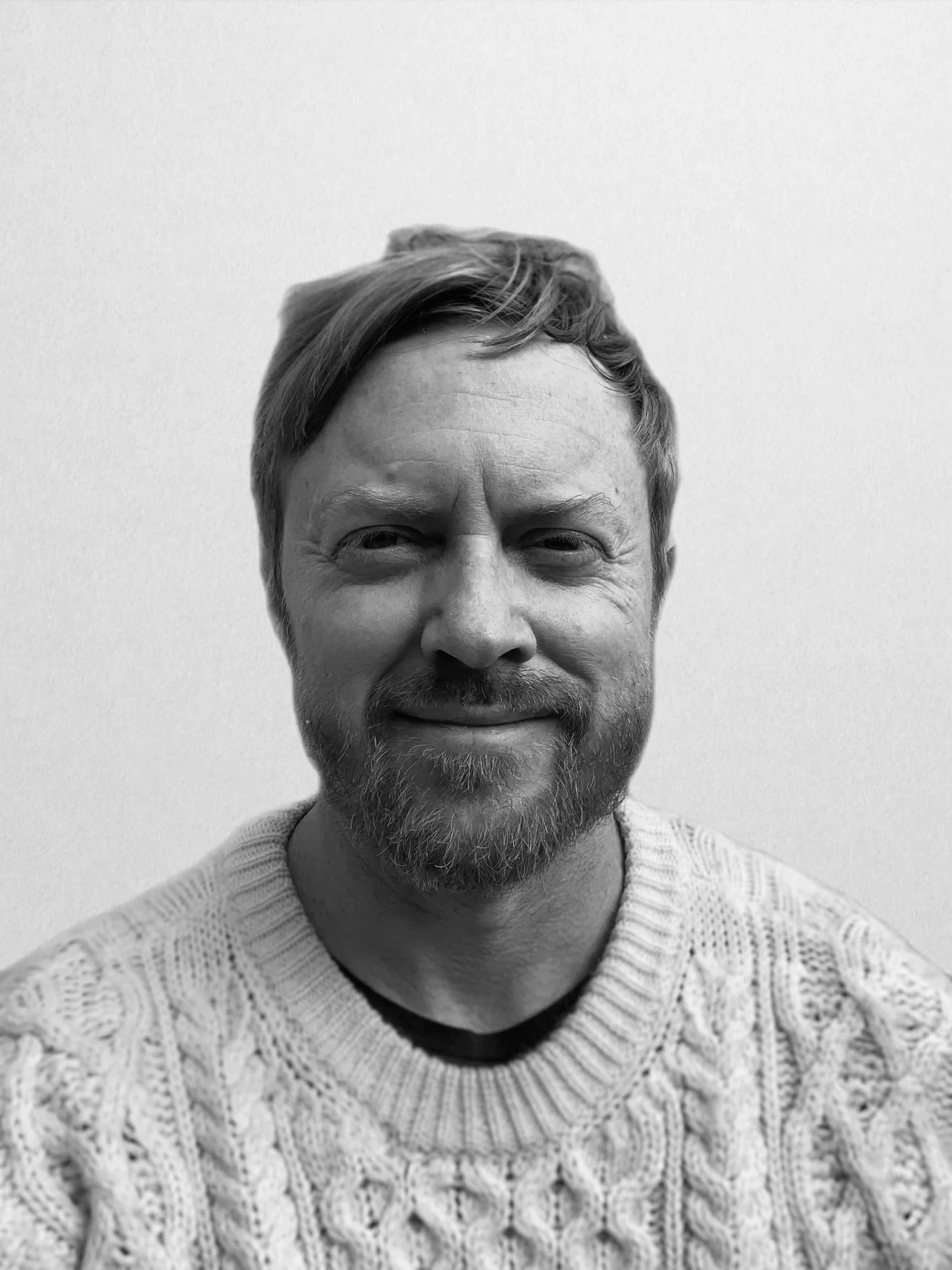Black and white portrait of a smiling man with light hair, beard, and mustache, wearing a cable-knit sweater.