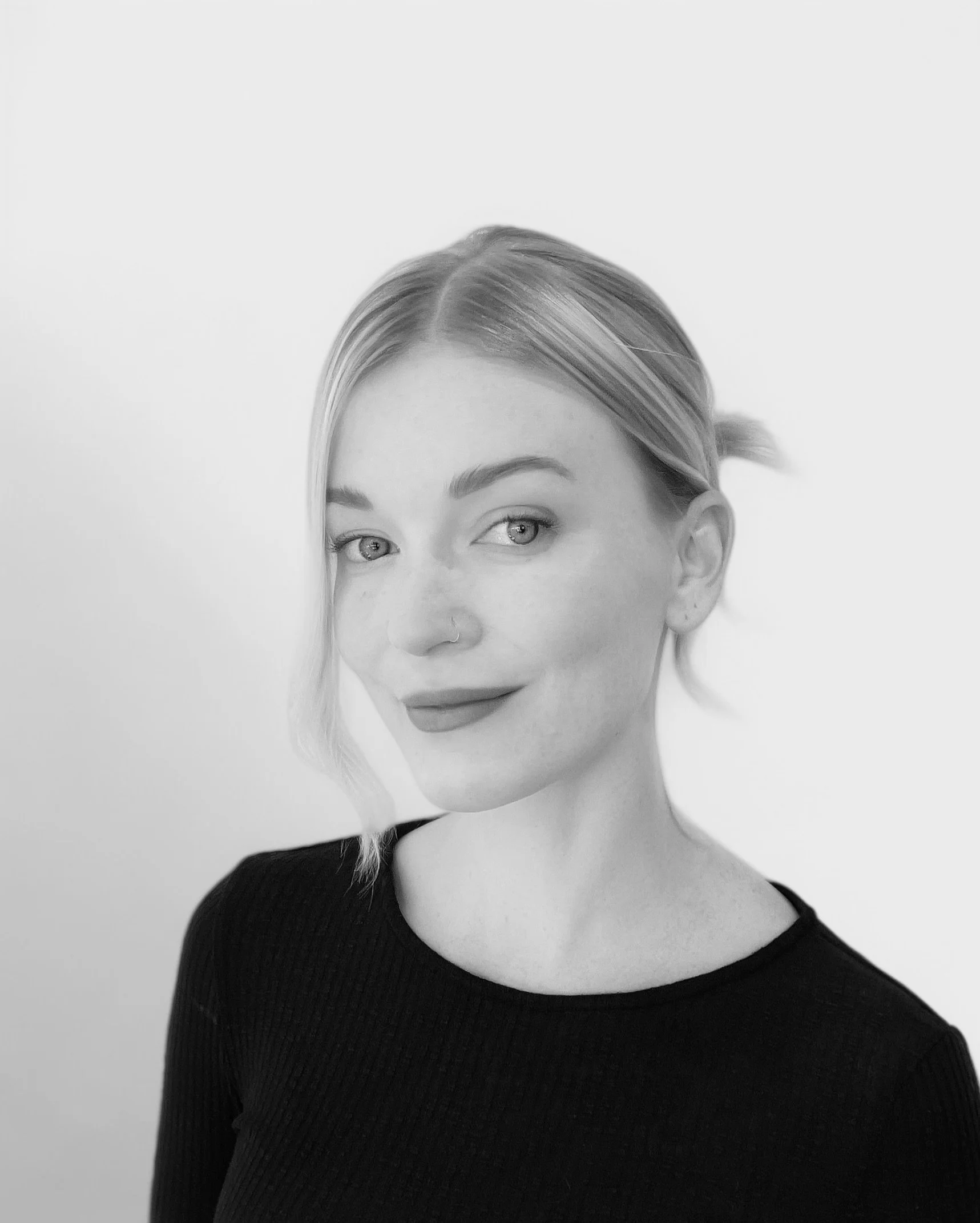 Black and white portrait of a young woman with light-colored hair, styled with a small side bump, wearing a dark ribbed top, smiling subtly with a confident expression, set against a plain background.
