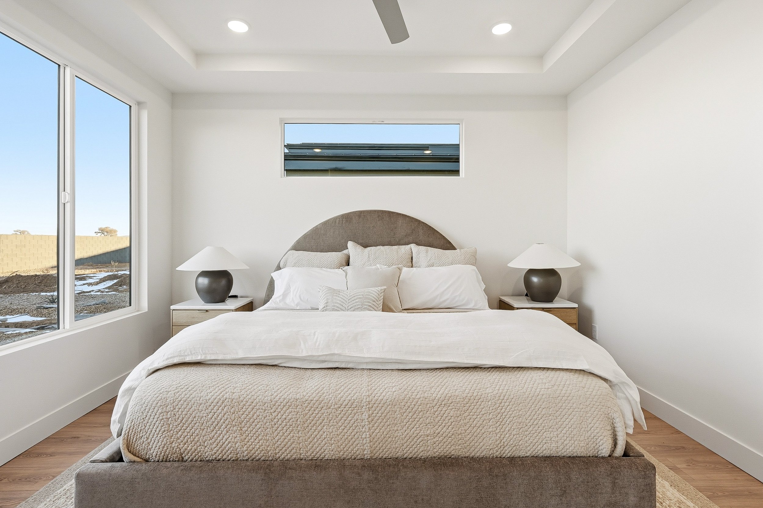 Minimalist bedroom with neutral tones, featuring a bed with beige bedding, two bedside tables with lamps, three wall pictures, a window with curtains, a chair, and a small side table.