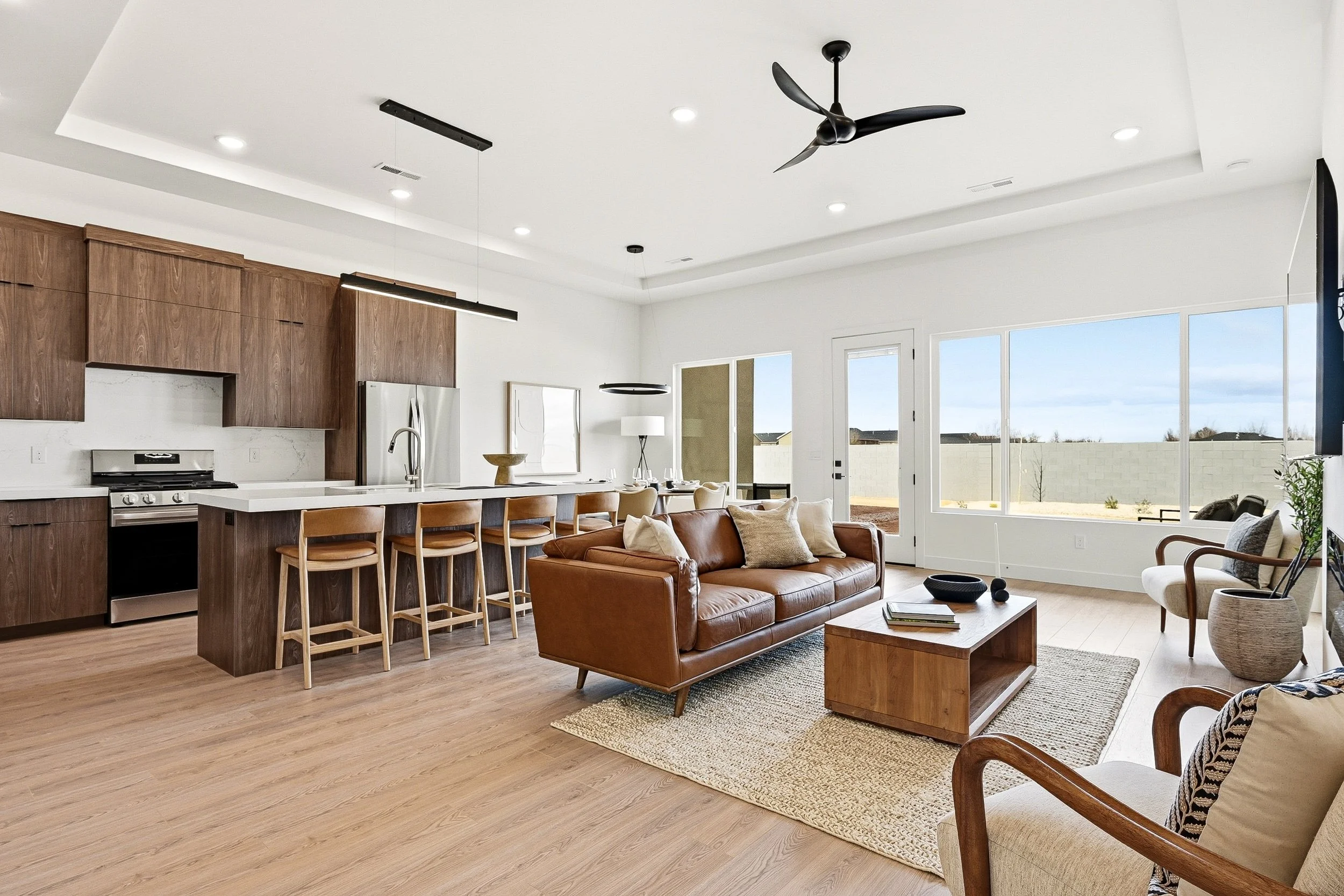 Open-concept living room and kitchen with wood flooring, large windows, modern ceiling fan, and seating area.