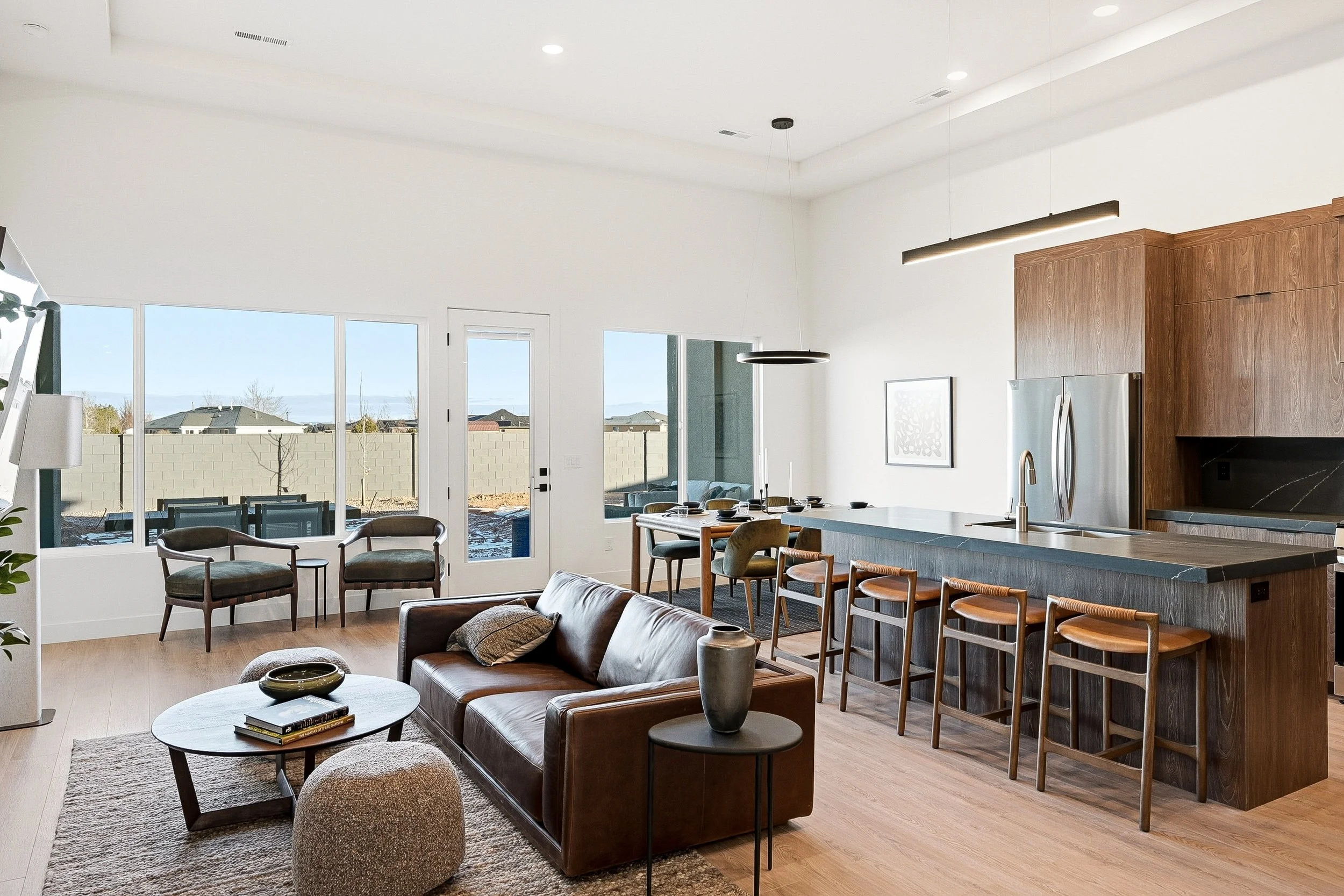 Modern open-concept living room and kitchen with white cabinetry, a kitchen island, a gray sofa, a wooden dining table with black chairs, and large windows allowing natural light.