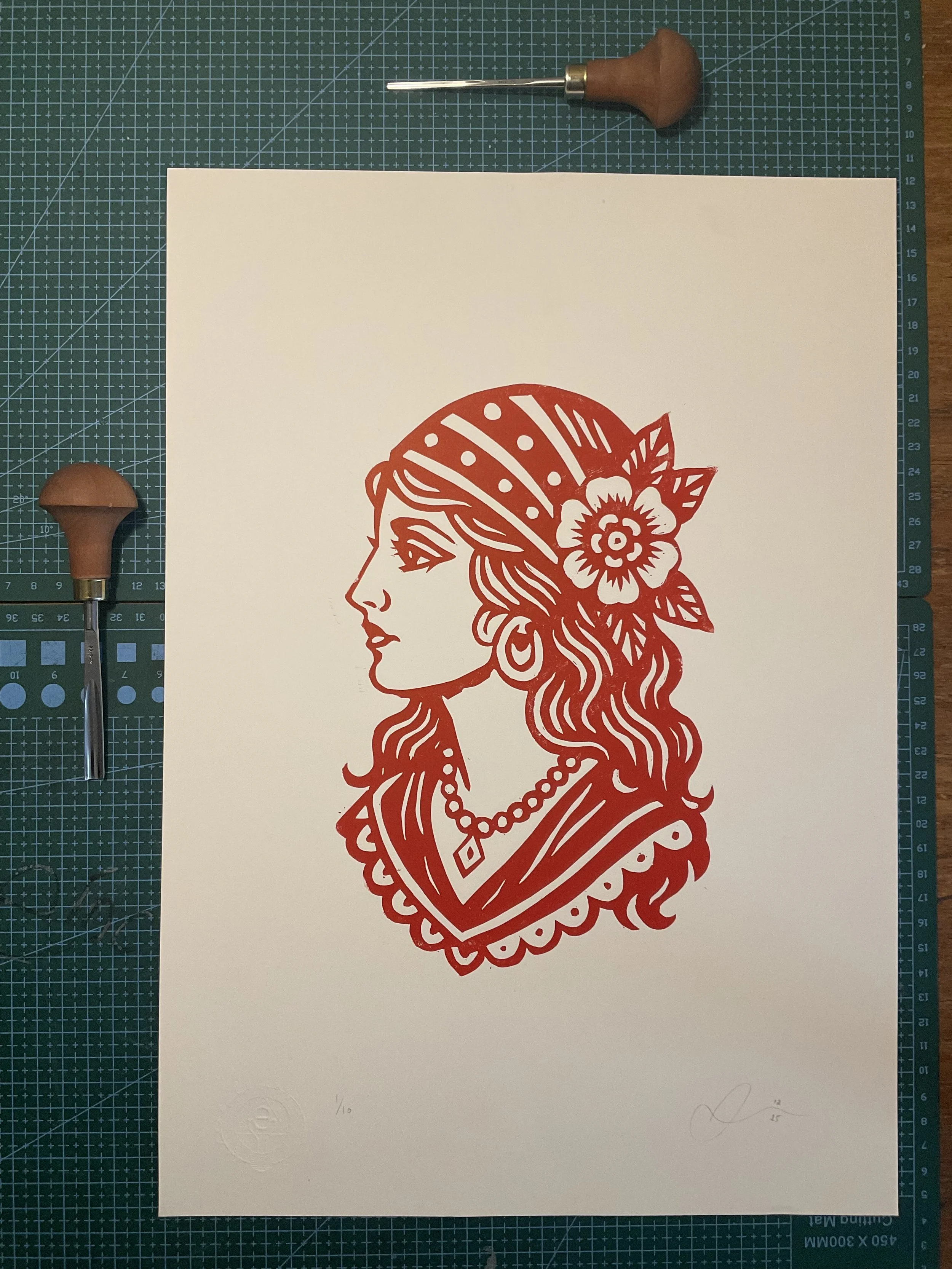 Red linocut print of a traditional tattoo-style gypsy woman with headscarf, flower and jewellery. Hand-carved and hand-printed by Elucidation Inc.”