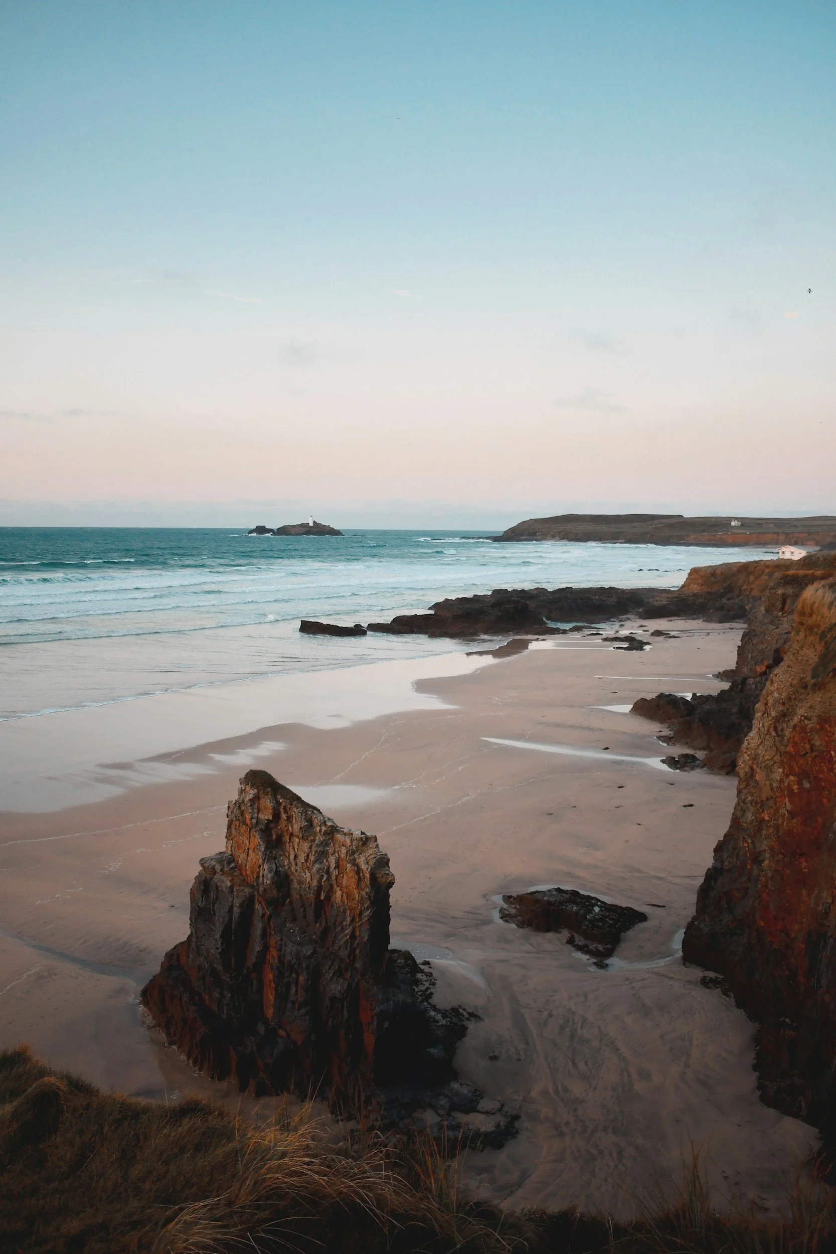 Coastal scene with rocky shoreline, sandy beach, ocean waves, and distant lighthouse on small island during sunset or sunrise.