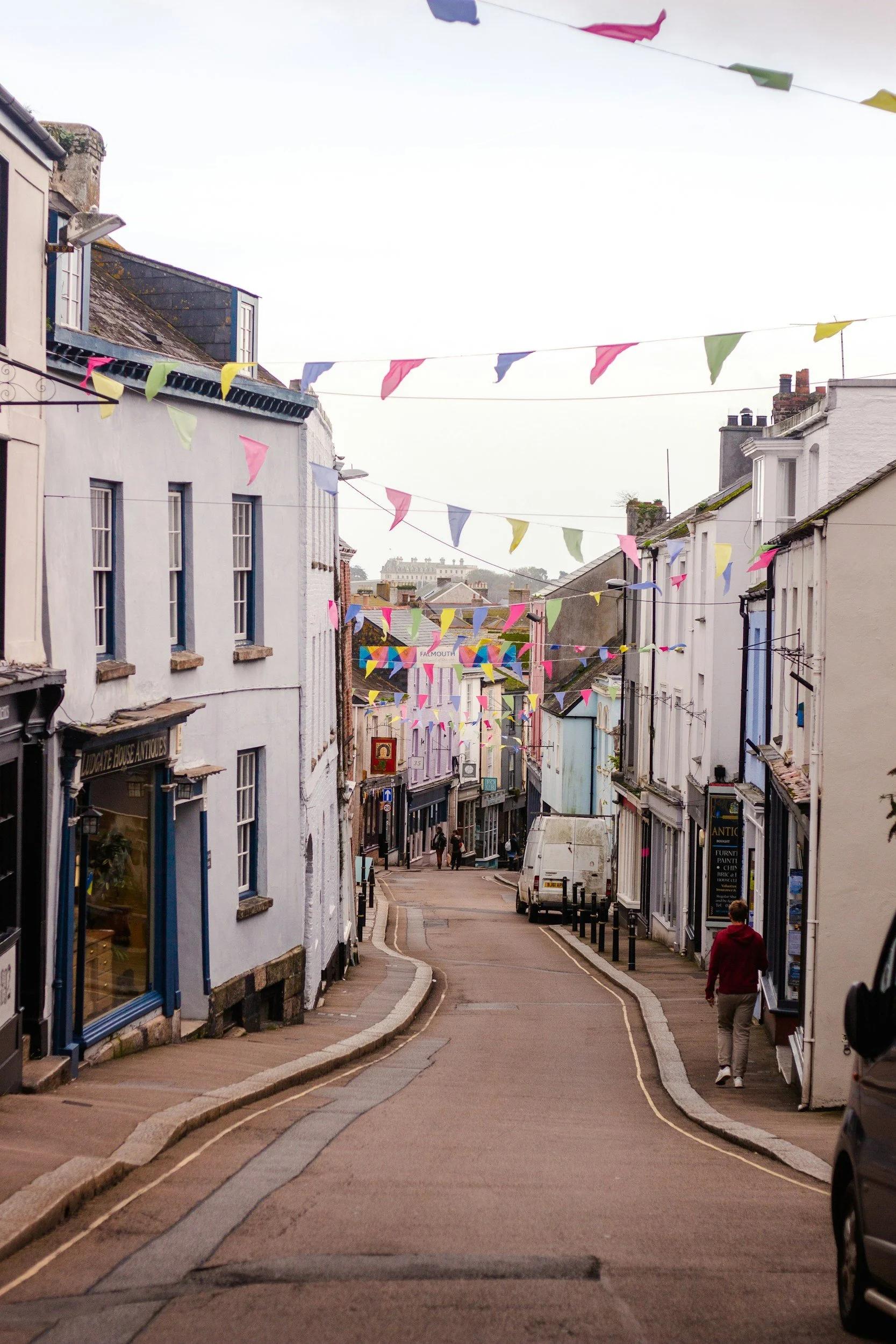 A street scene with colorful bunting flags hanging overhead, residential buildings on either side, and a few people walking on the sidewalk. A white van is parked on the right, and a person wearing a red hoodie is walking away from the camera.