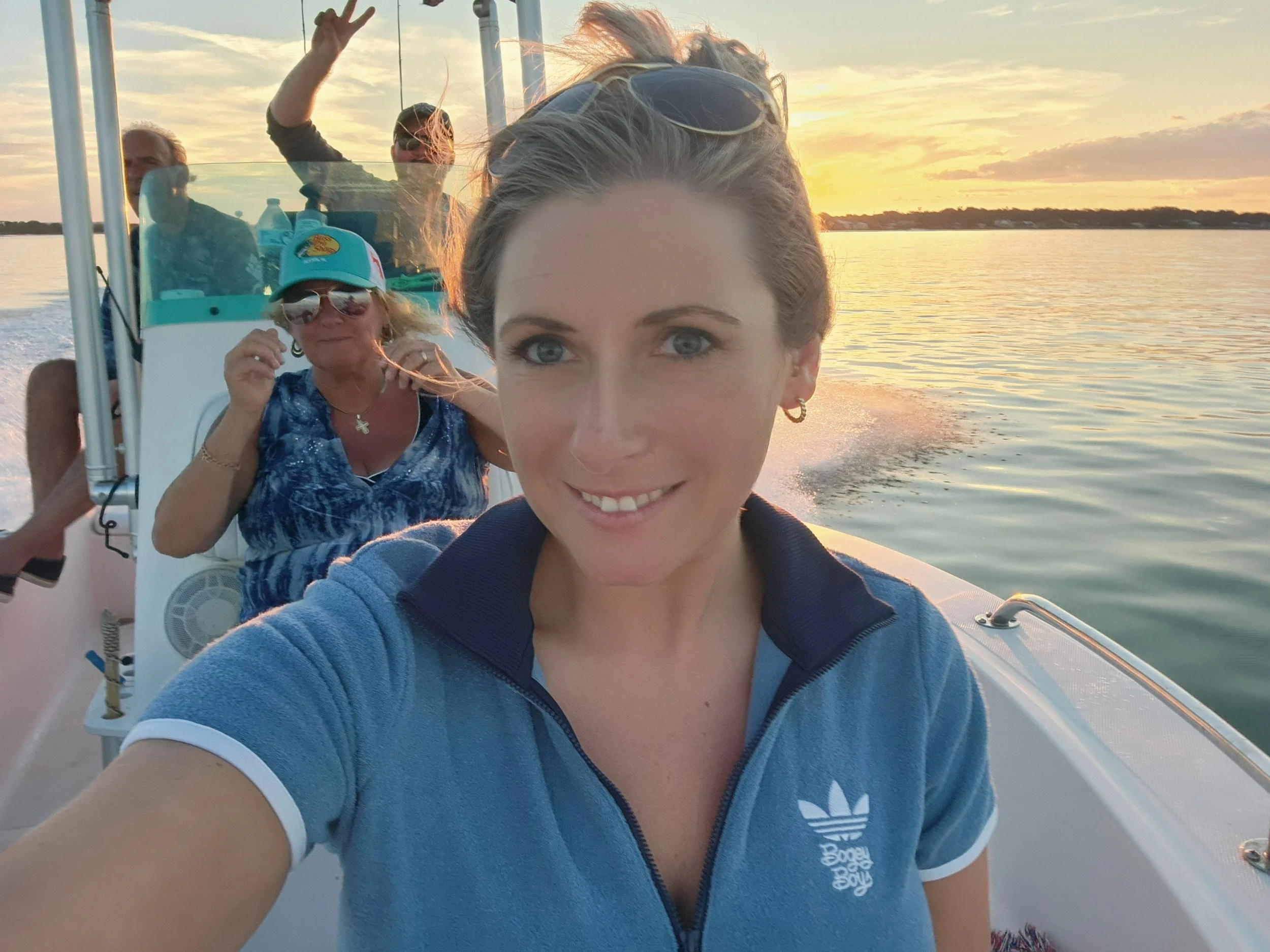 A group of four people on a boat during sunset on a calm body of water. The young woman in the foreground is smiling at the camera, wearing a blue Adidas jacket and sunglasses on her head. Behind her, an older woman with curly hair, wearing sunglasses, a hat with a fish logo, and a blue patterned shirt, is giving a thumbs-up. Two men in the background are near the boat's console, one making a peace sign with fingers, both enjoying the sunset atmosphere.