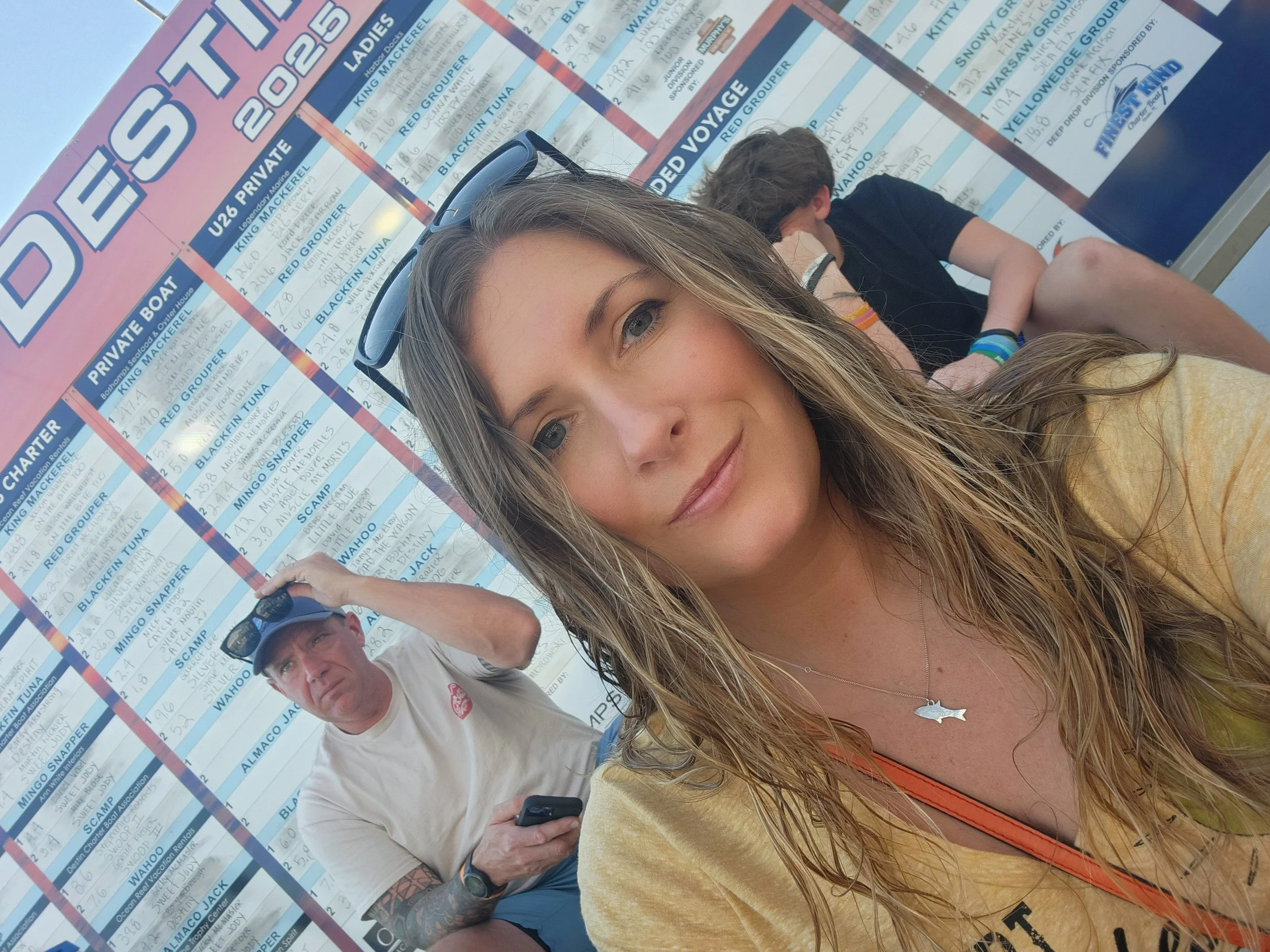 A young woman with long blonde hair, wearing a yellow shirt and a fish necklace, smiling at the camera, standing in front of a large boat schedule sign. Behind her, a man in a white shirt and black cap is looking at his phone.