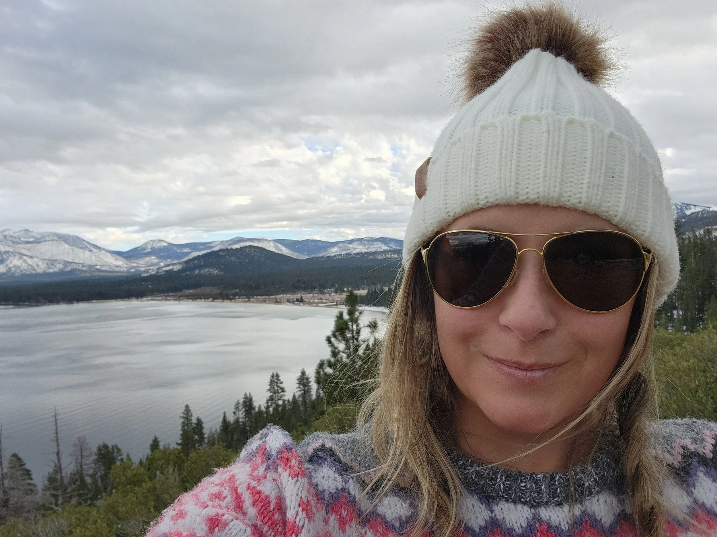 A woman wearing sunglasses, a white knit beanie with a brown pom-pom, and a colorful sweater standing outdoors with mountains, trees, and a lake in the background.