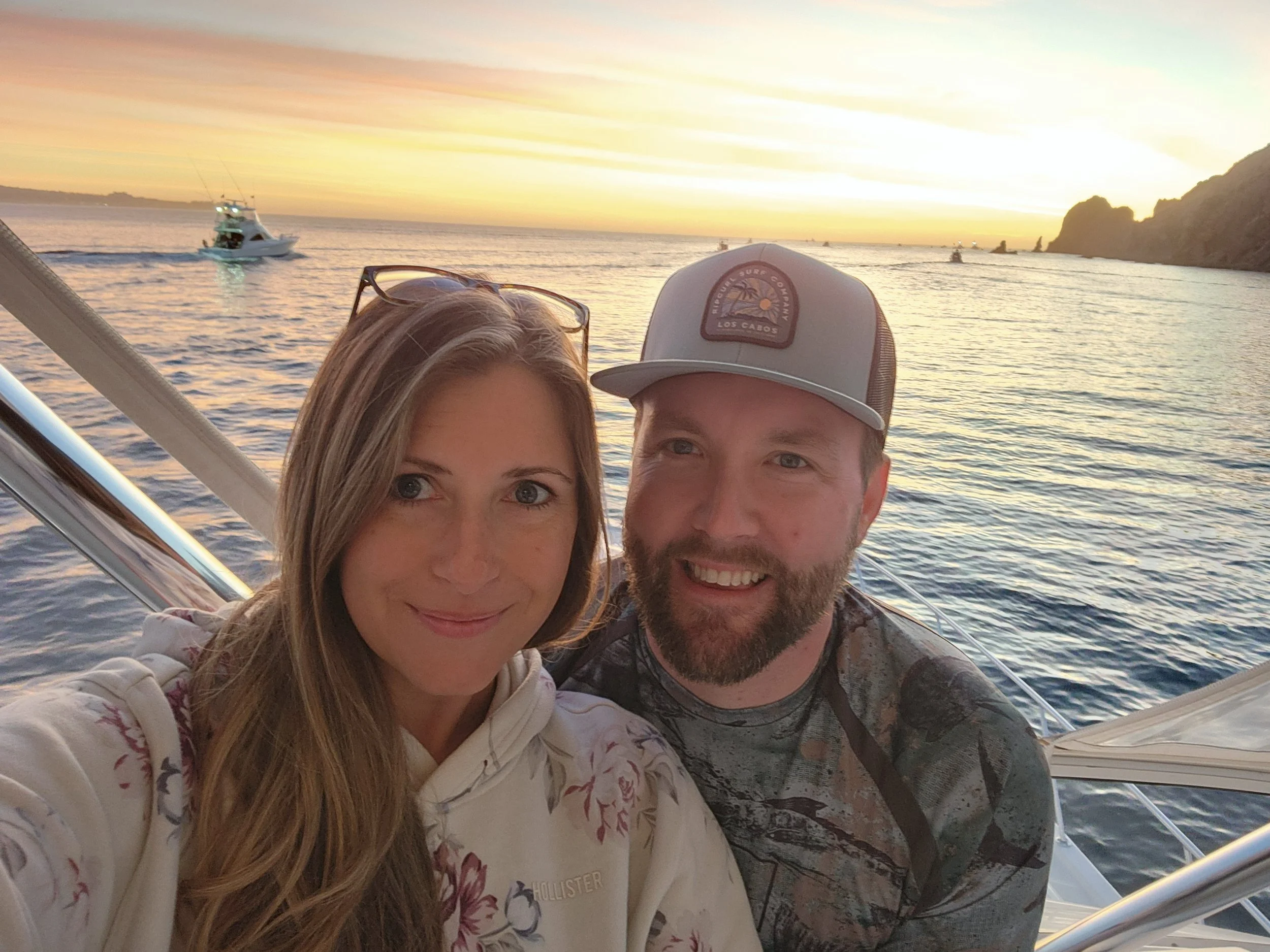 A couple taking a selfie on a boat with a sunset view over the ocean, with boats and rocky cliffs visible in the background.