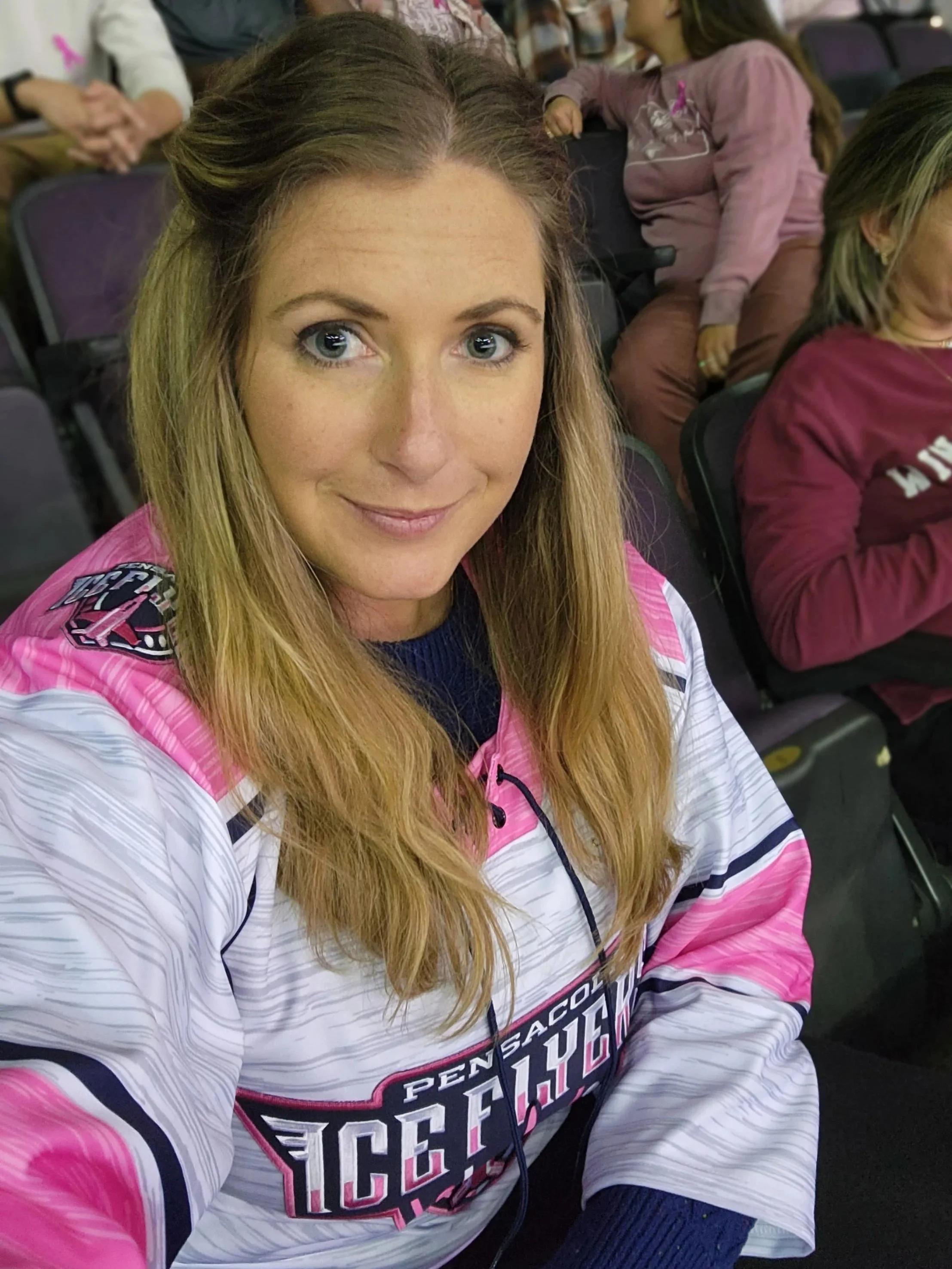 A woman with long blonde hair and blue eyes wearing a racing team jersey is taking a selfie in a room with other people seated around her.
