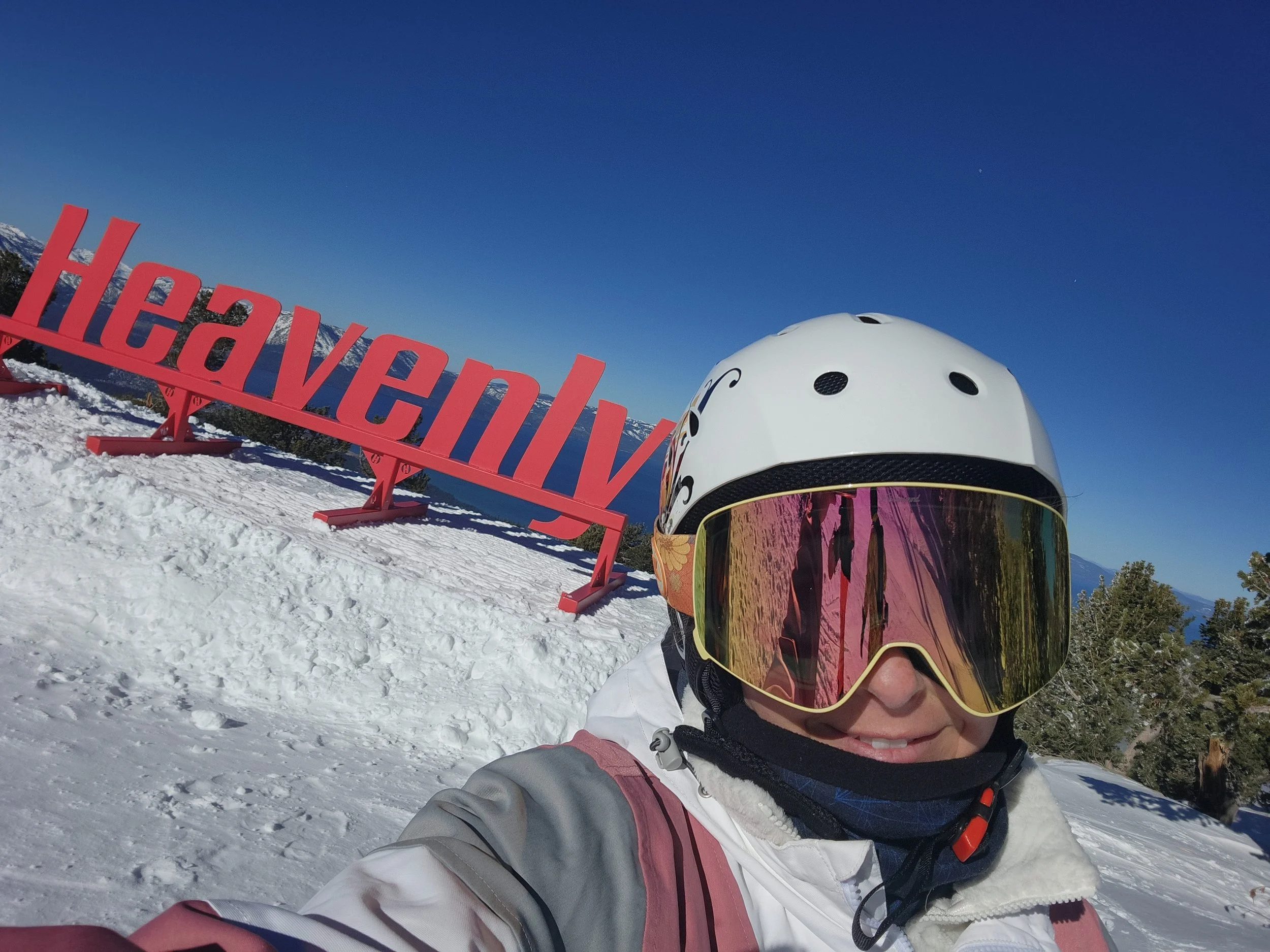 A person in ski gear with a white helmet and goggles taking a selfie on a snow-covered mountain during daytime. A red sign reading 'Heavenly' is visible in the background along with trees and clear blue sky.