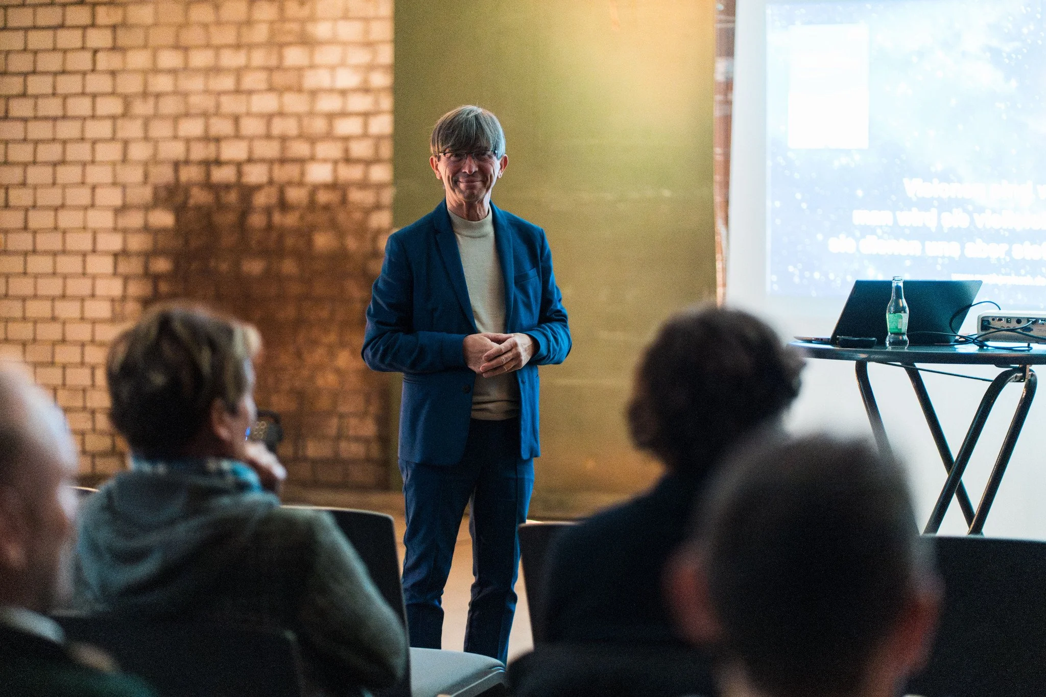 Mann hält einen Vortrag vor einer Gruppe von Zuhörern in einem Raum mit einer Projektionsleinwand.