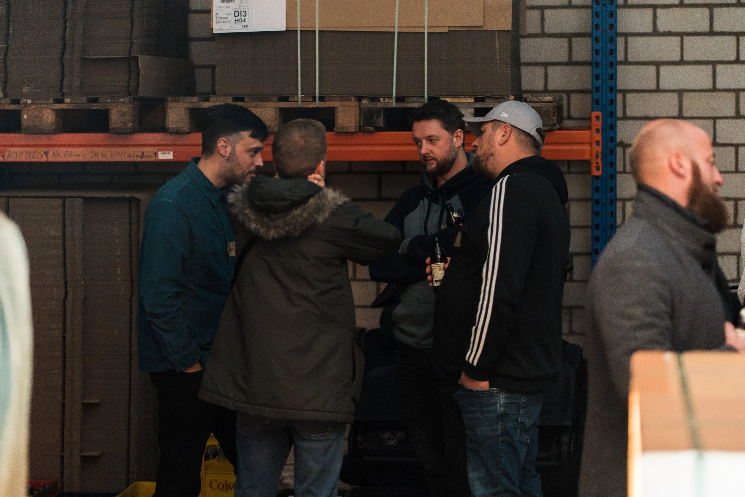 Fünf Männer stehen in einem Lagerraum, einige halten Bierflaschen und sprechen miteinander.