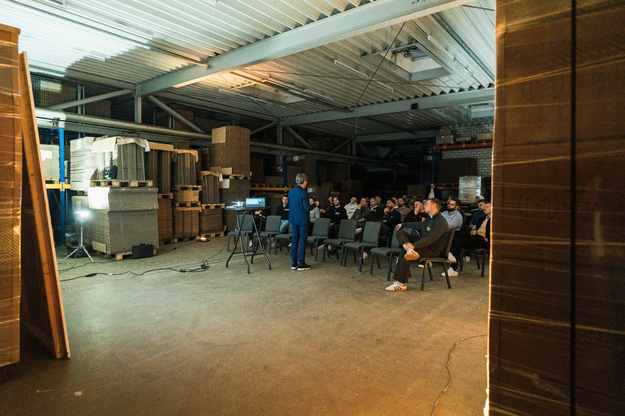 Eine Gruppe von Menschen sitzt in einem Industriegebäude in einem Seminar oder Vortrag. Ein Mann, der wahrscheinlich der Referent ist, steht vor der Gruppe und spricht. Es gibt Stühle, einen Laptop auf einem Tisch, und das Gebäude ist mit Lagerregale