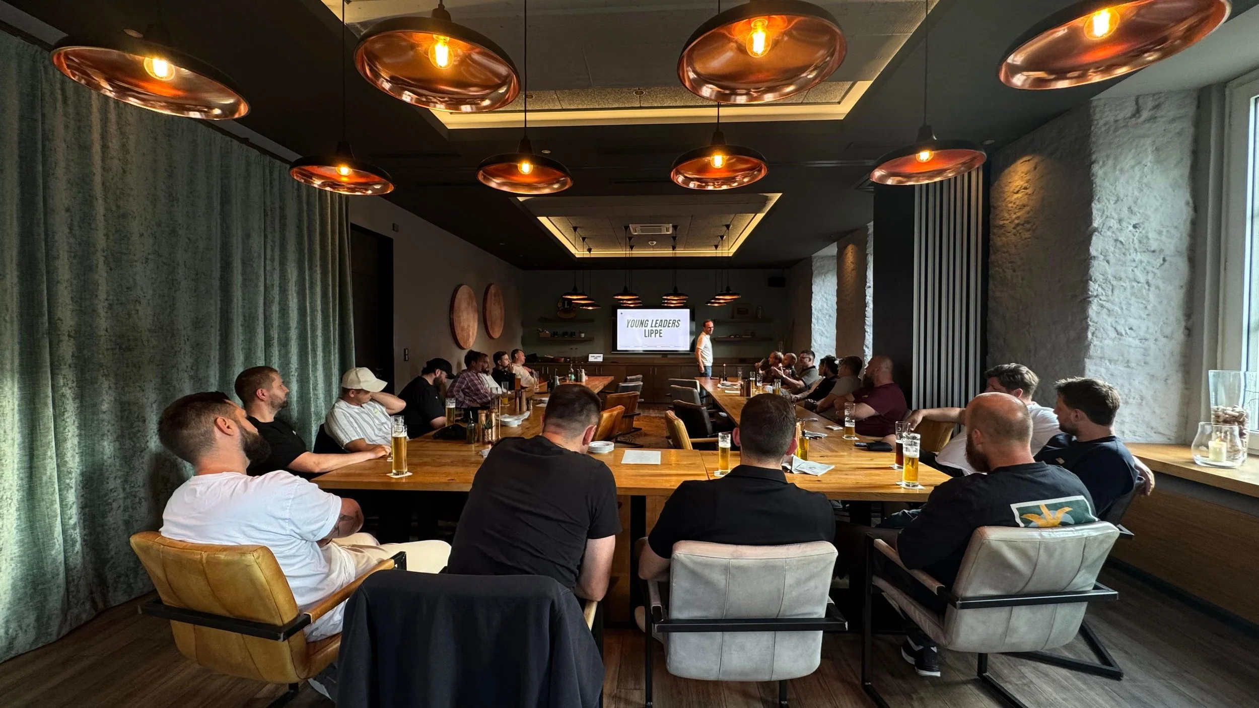 Ein Meetingraum mit einer großen runden Tafel, um die herum Menschen sitzen, die einem Vortrag folgen. Im Hintergrund ist ein Bildschirm mit der Aufschrift 'Young Leaders Lippe'. Der Raum ist modern gestaltet mit Kupferpendeln an der Decke und großen