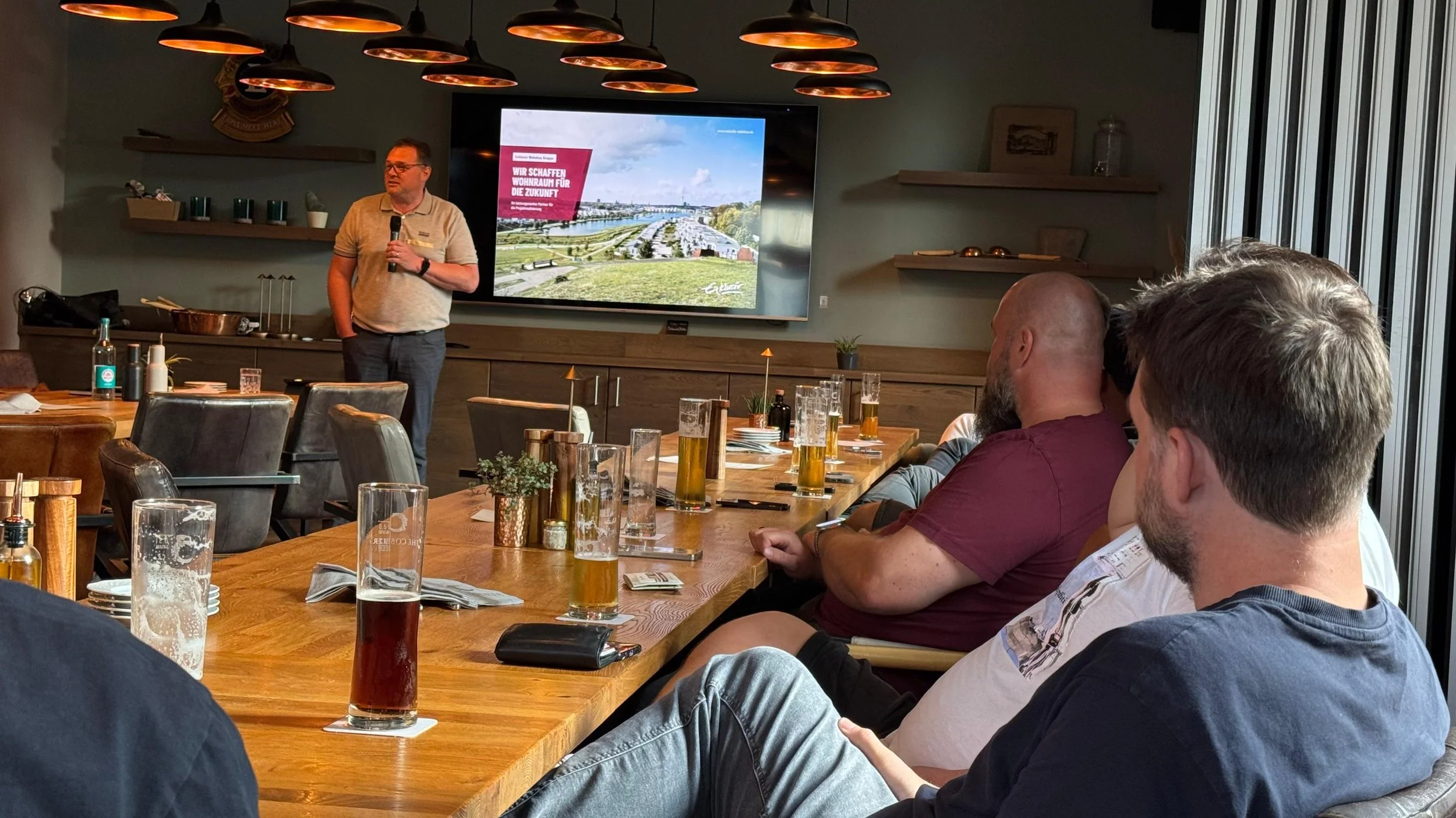 Ein Mann hält eine Präsentation vor einem Raum voller Menschen, die an einem langen Holztisch sitzen. Auf dem Bildschirm im Hintergrund ist eine Präsentationsfolie mit einem Bild und deutschem Text.”}}