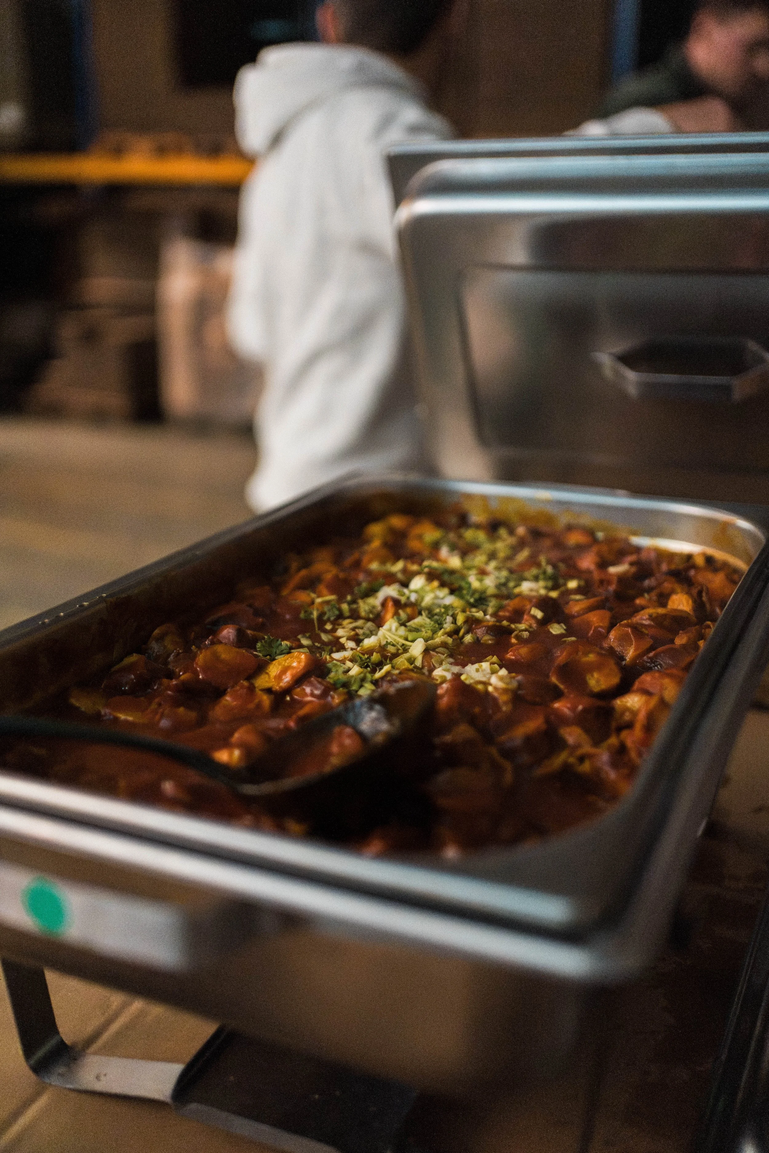 Ein chafiertes Gericht in einer Edelstahl-Servierpfanne mit gehacktem Grün als Garnitur, im Hintergrund sind zwei Personen in einem Restaurant oder Café.