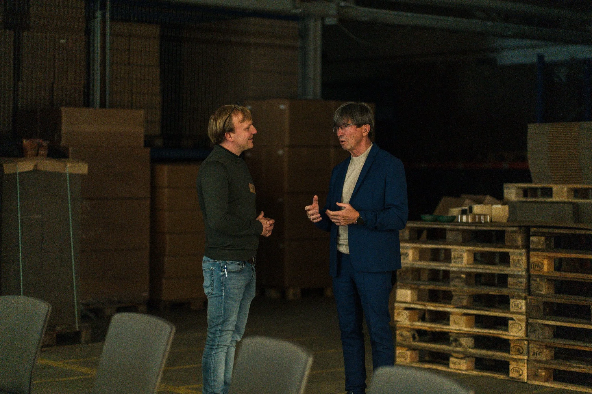 Zwei Männer unterhalten sich in einem Lagerraum mit Paletten und großen Kisten.