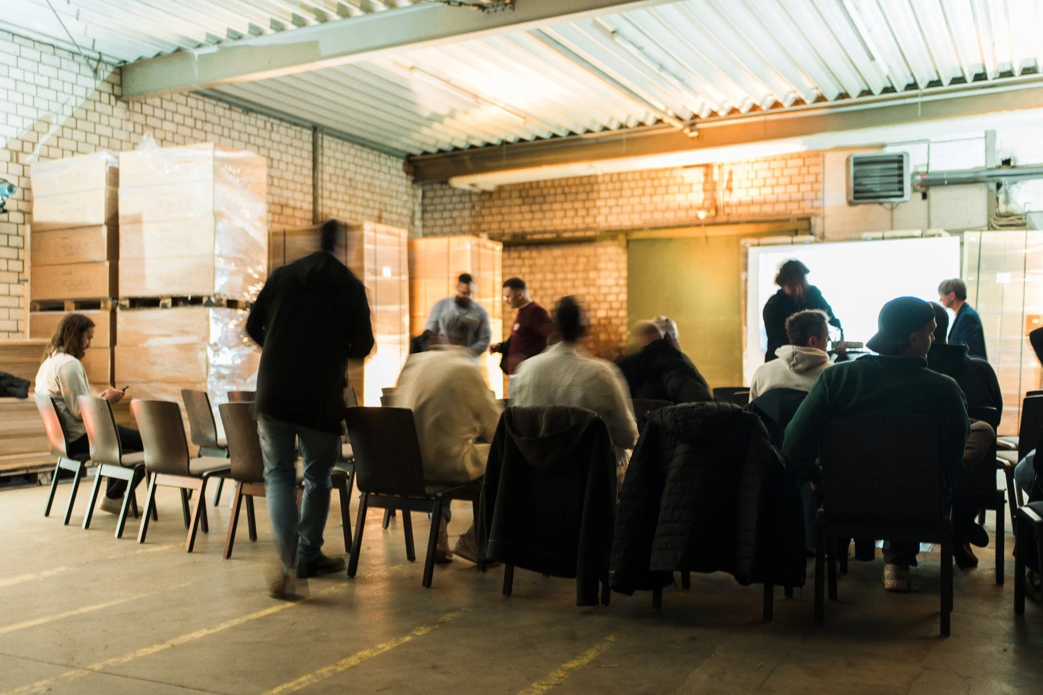 Menschen sitzen und stehen in einem industriellen Raum mit Backsteinwänden, gestapelten Kartons und einem Projektor oder Bildschirm an der Wand, beim Treffen oder Workshop.