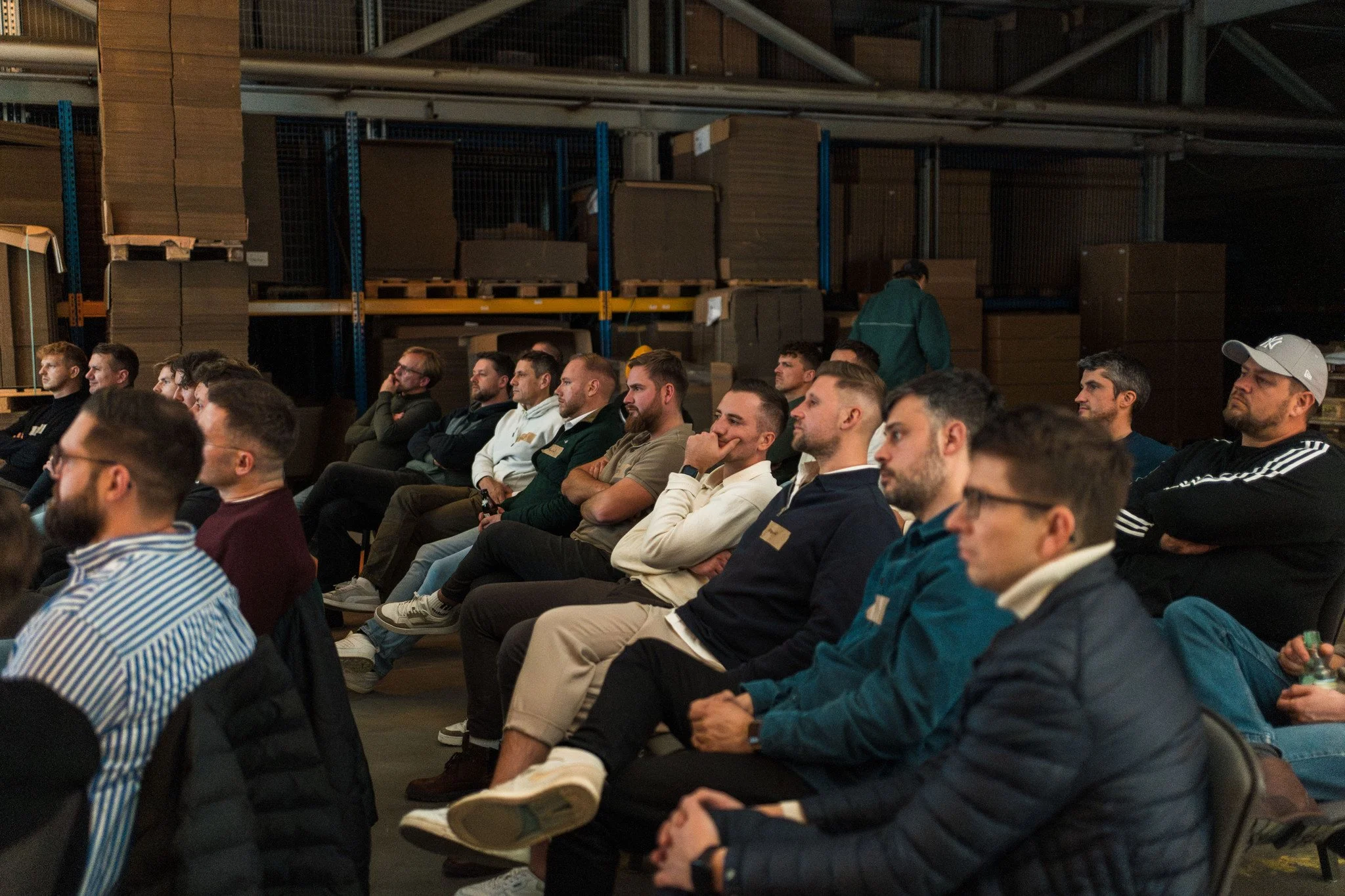Menschen sitzen in einer Reihe bei einer Konferenz in einer Lagerhalle oder Werkhalle, im Hintergrund sind Regale mit Kartons und Holzwaren.