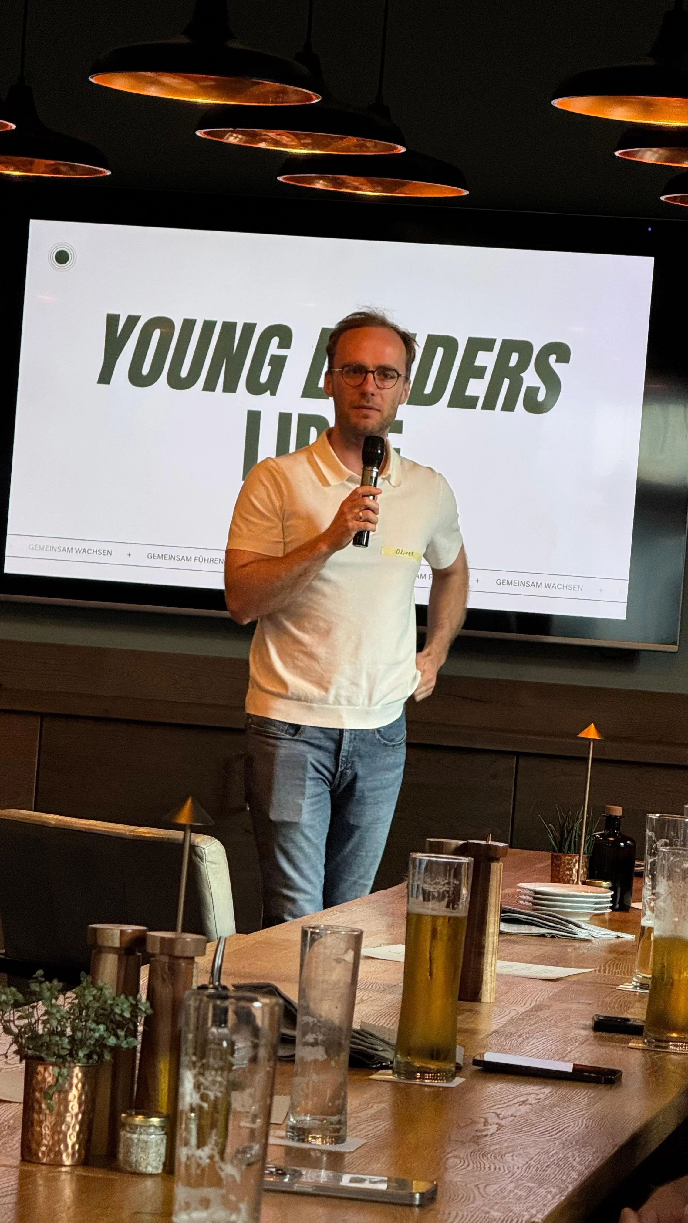 Ein Mann hält ein Mikrofon beim Vortrag vor einer großen Bildschirm mit der Aufschrift "Young Leaders". Im Vordergrund sind Tische mit Gläsern und Dekorationen zu sehen.