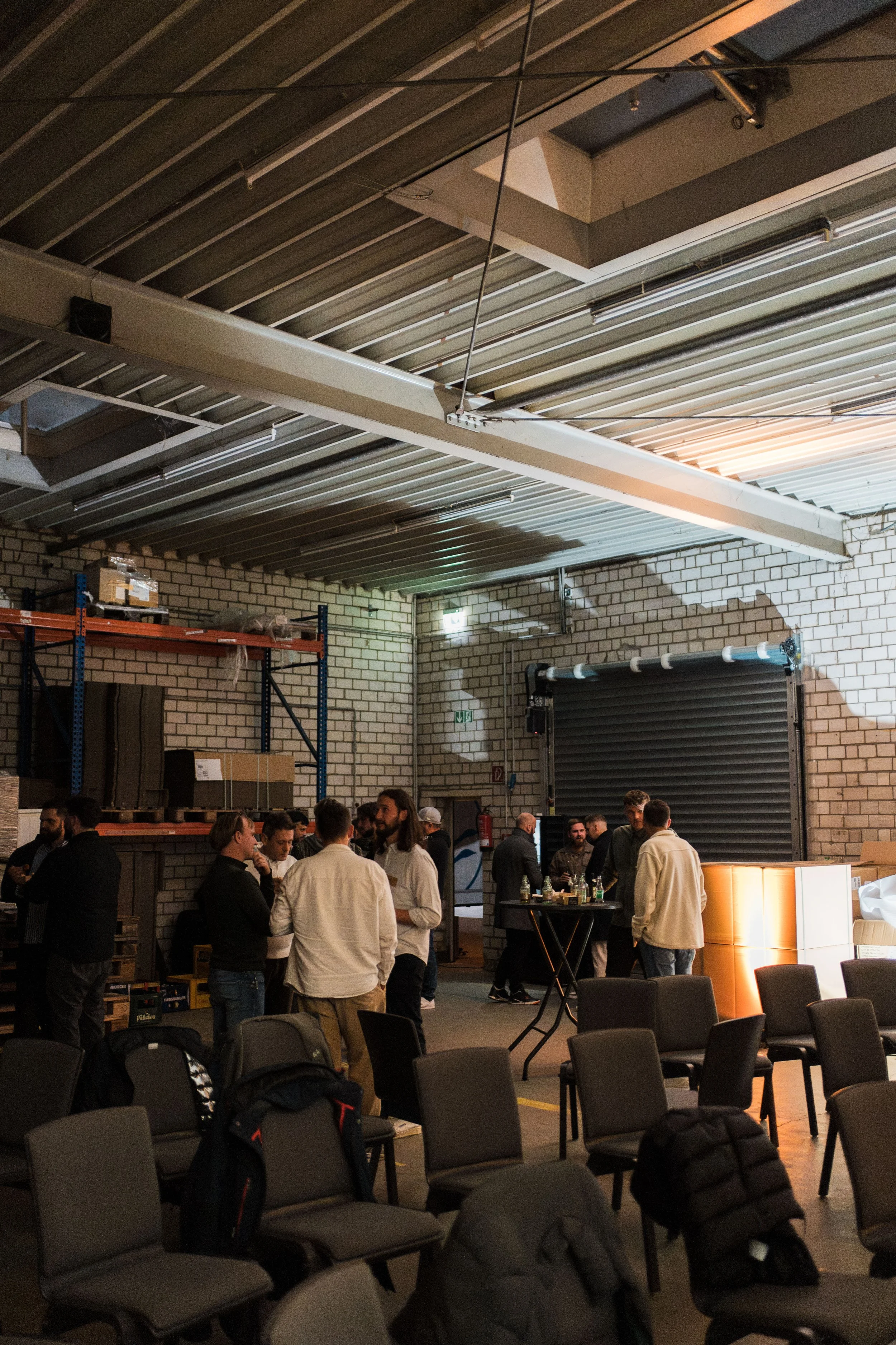 Menschen stehen in einer Lagerhalle mit weißen Ziegelwänden. Es gibt Tische, Stühle und Hochregale mit Kartons. Eine Gruppe unterhält sich bei einem Getränk, im Hintergrund ein rolltor und Lagerregale.