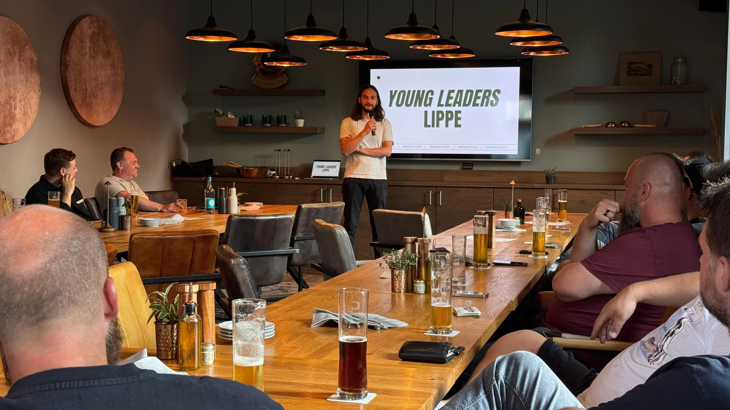 Ein Mann hält eine Präsentation vor einem Publikum in einem modernen Raum mit Holztisch und Fenstern, auf dem Bildschirm steht ‚YOUNG LEADERS LIMPE‘, Getränke auf dem Tisch, mehrere Zuhörer schauen zu.