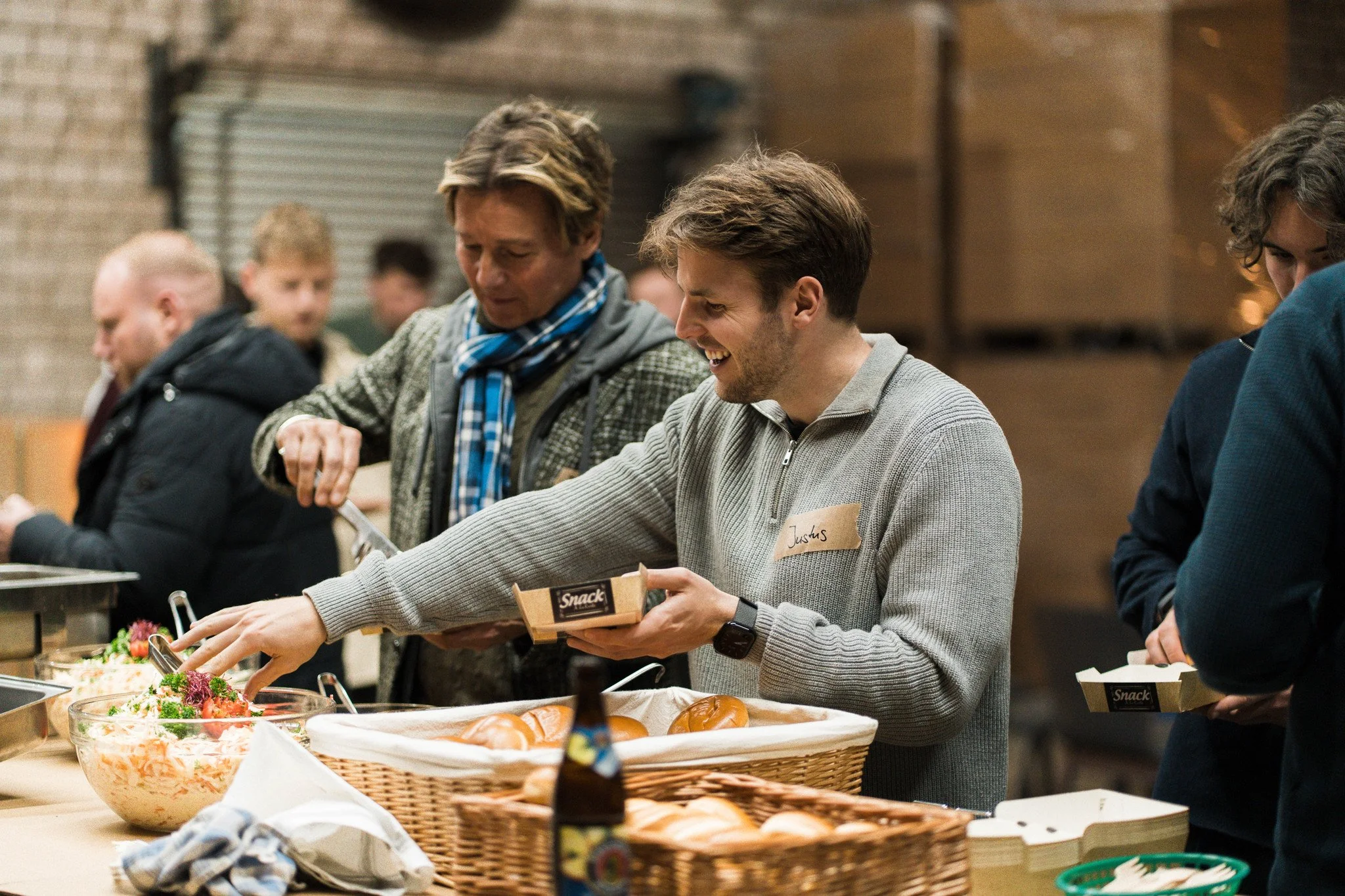 Menschen stehen in einer Warteschlange an einem Buffet mit Salat, Brötchen und Brot, in einer modernen, industriellen Umgebung.
