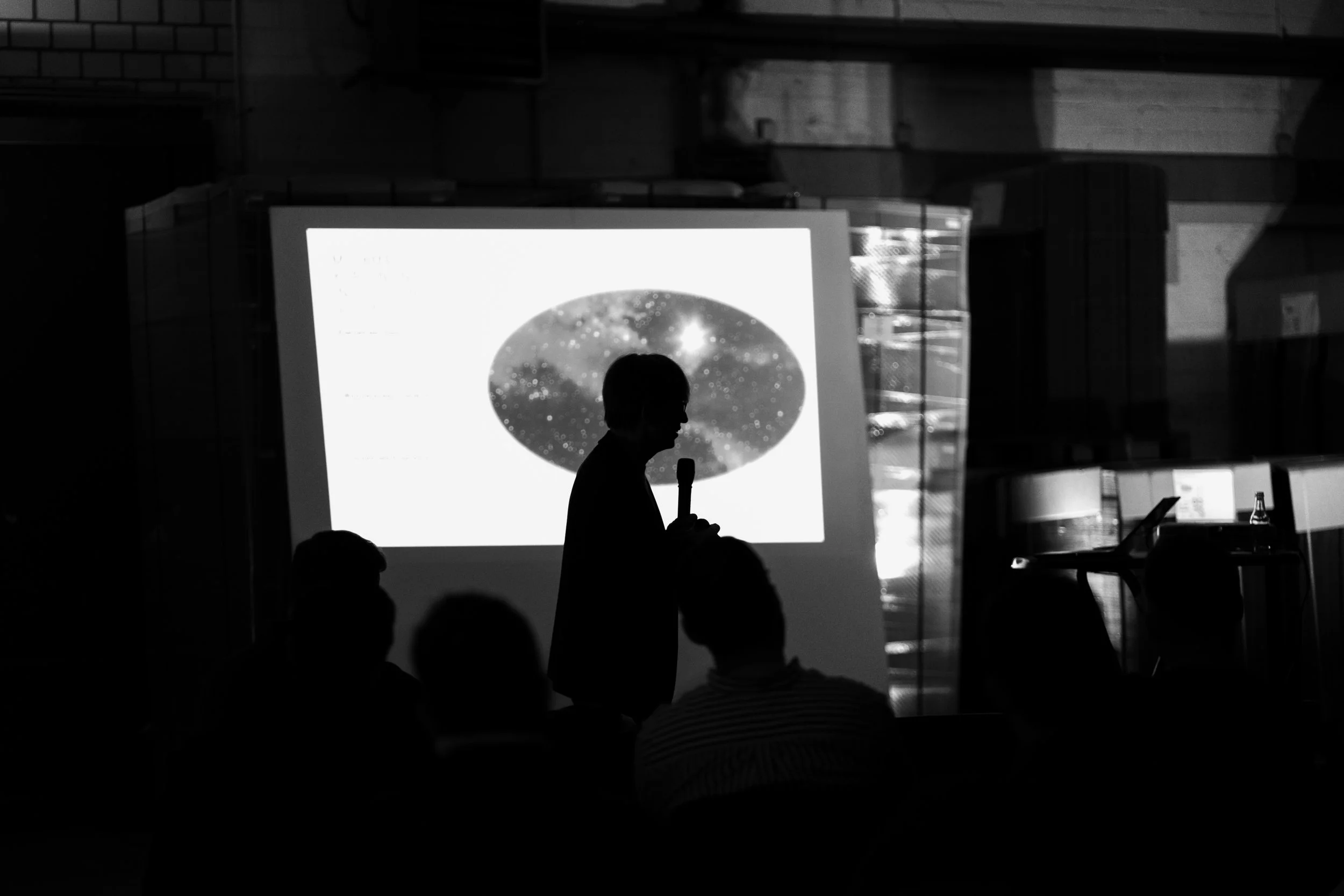 Eine Person hält ein Mikrofon und präsentiert vor einer Projektionswand, auf der ein Bild des Nachthimmels mit Sternen und Galaxien zu sehen ist. Das Bild ist in Schwarzweiß.