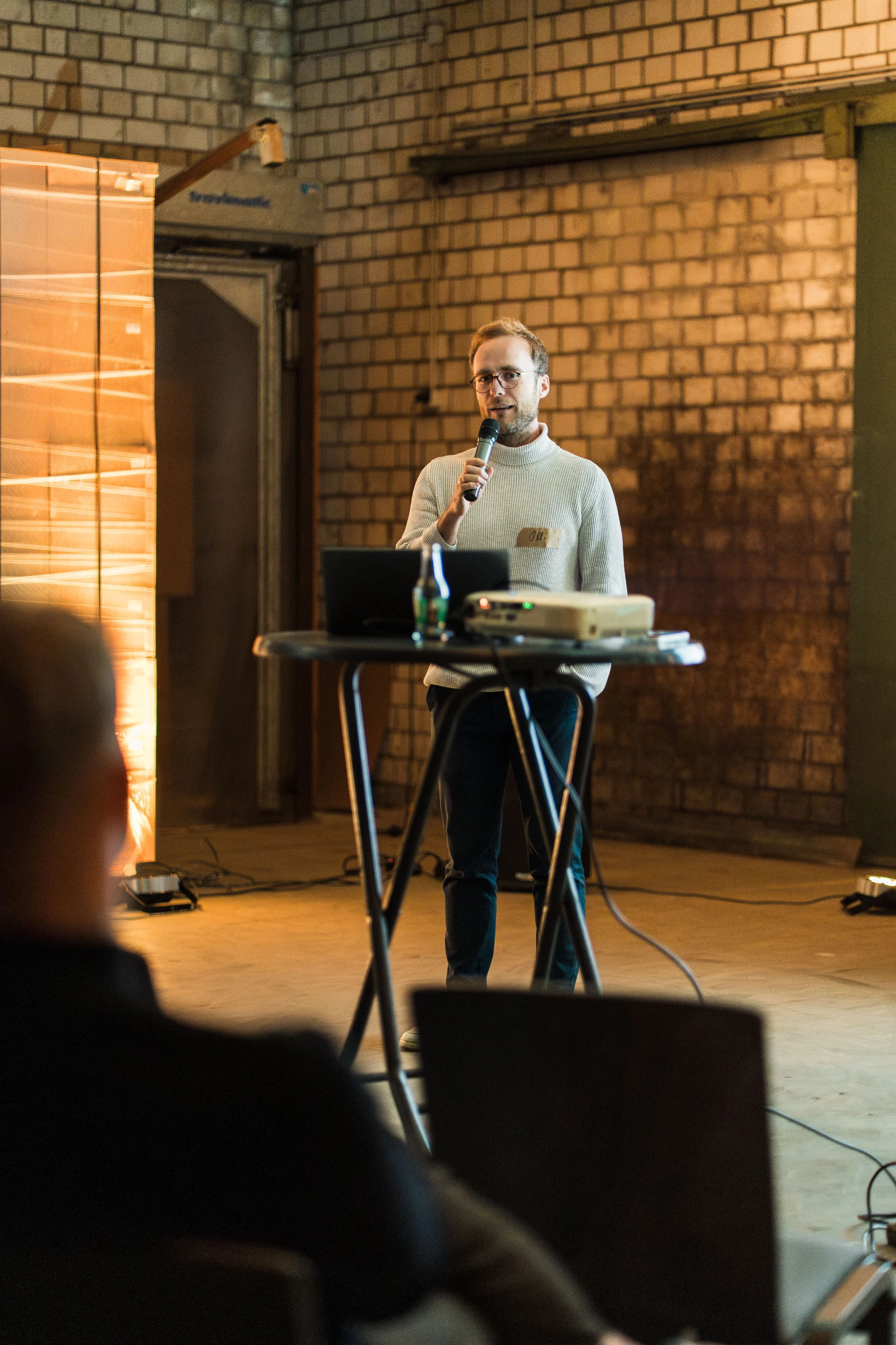 Ein Mann hält eine Präsentation vor einem Publikum in einem Raum mit Backsteinwänden, während er in ein Mikrofon spricht.