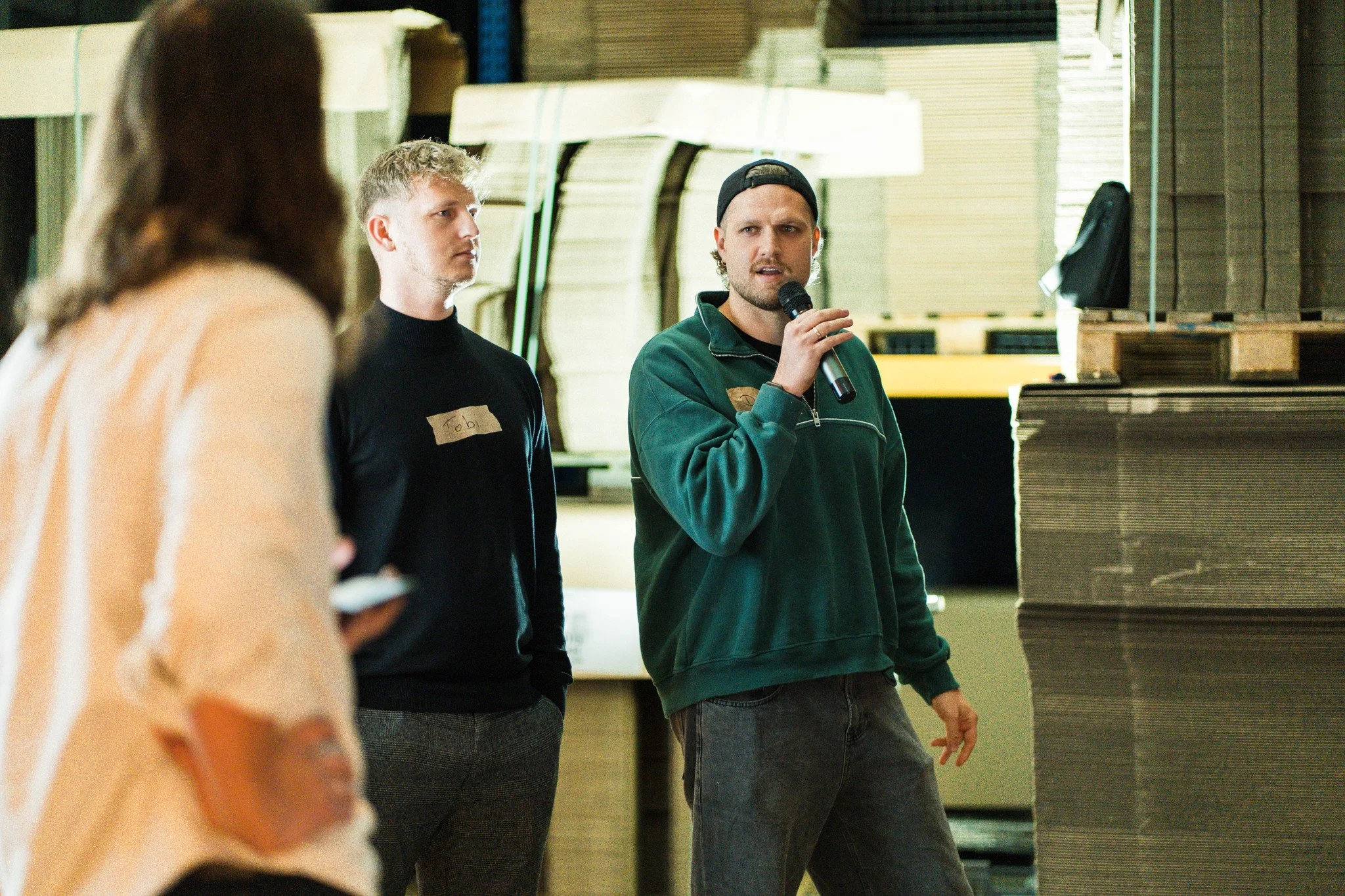 Drei Personen in einem Lager oder Fabrik, eine spricht mit Mikrofon, die anderen hören zu.