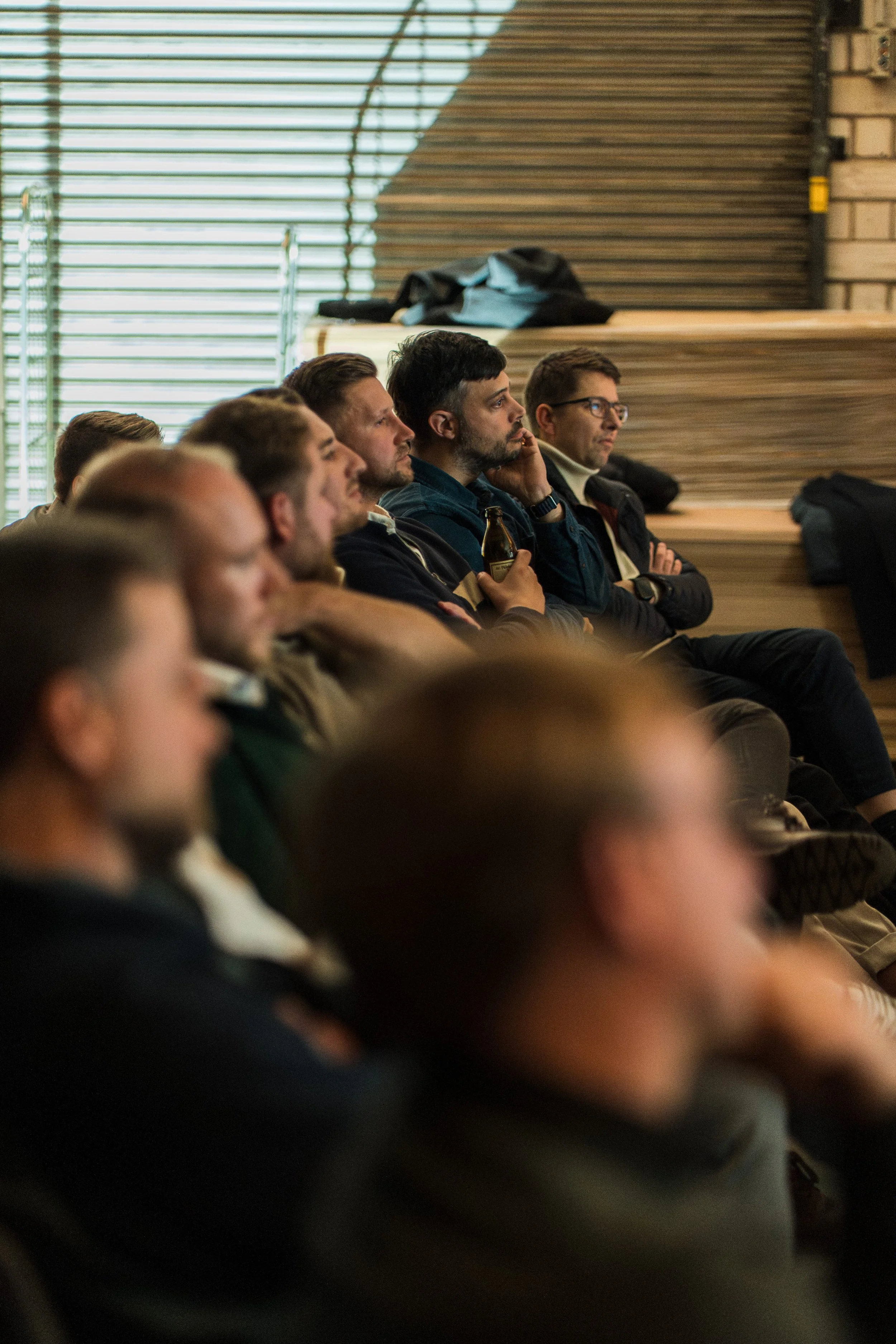 Menschen sitzen in einer Reihe und schauen aufmerksam eine Präsentation oder einen Vortrag an, während sie in einem Raum mit Holzstufen und großen Fenstern im Hintergrund sitzen.