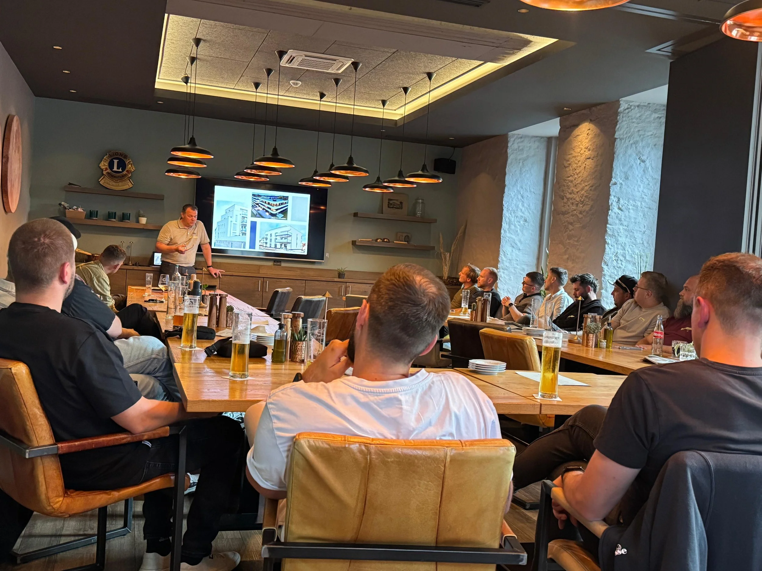 Ein Mann hält eine Präsentation vor einer Gruppe von Menschen in einem modernen Raum mit Holz- und Steinelementen, während er auf eine Wand mit Bildschirm zeigt.