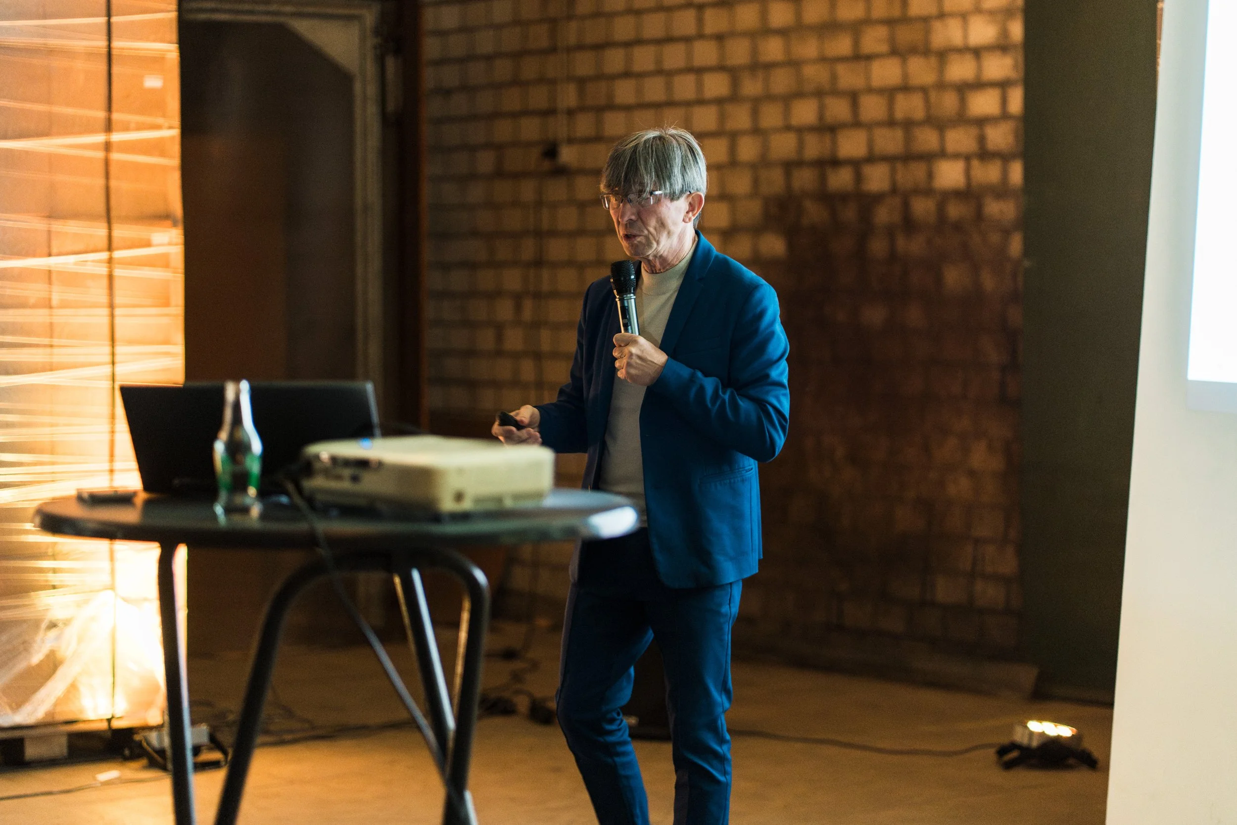 Ein junger Mann in blauer Anzug hält eine Mikrofon in der Hand und hält einen Vortrag in einem Raum mit Ziegelwänden.