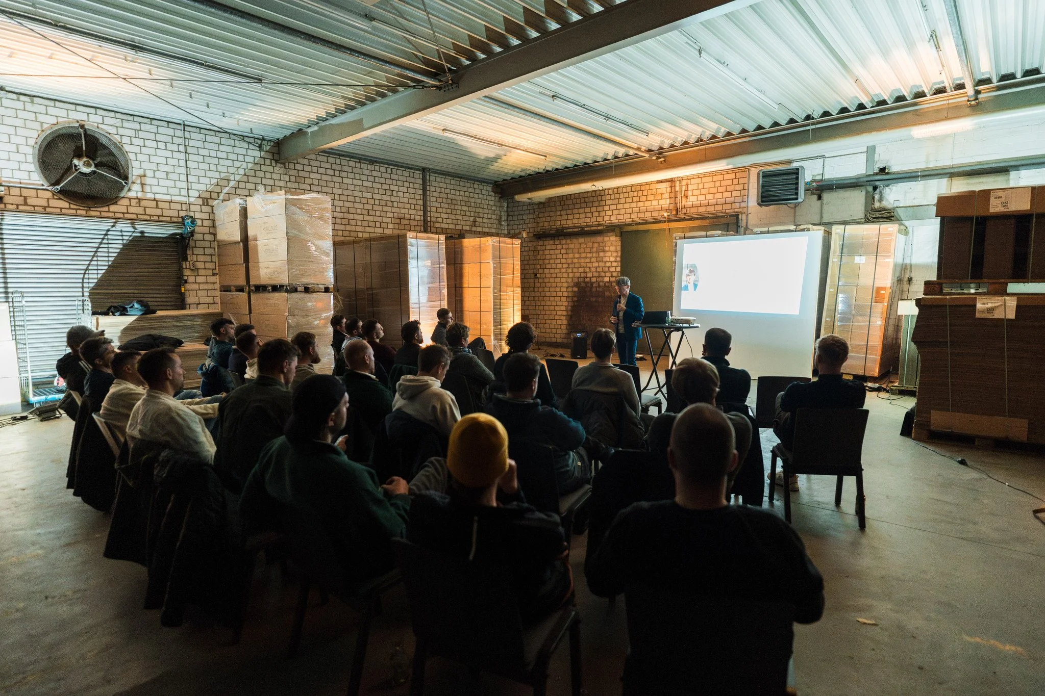 Eine Gruppe von Menschen sitzen in einem Raum mit Industrietechnik anwesend bei einer Präsentation, die von einem Sprecher vor einem großen Bildschirm gehalten wird.