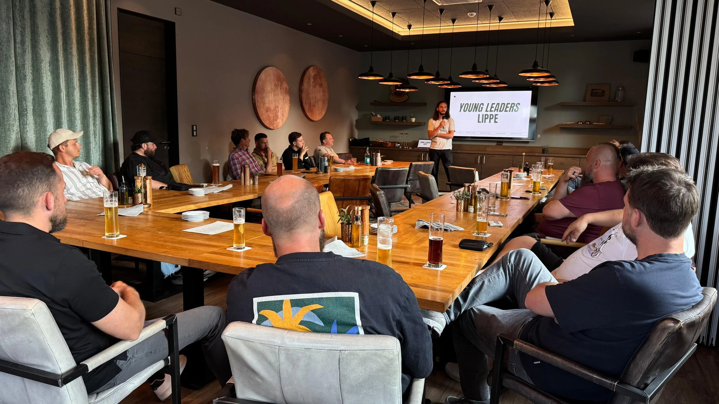 Eine Gruppe von Menschen sitzt an einem großen Holztisch in einem Konferenzraum und hört einem Vortragenden zu, der vor einem Bildschirm mit der Aufschrift "YOUNG LEADERS LIPPE" steht.