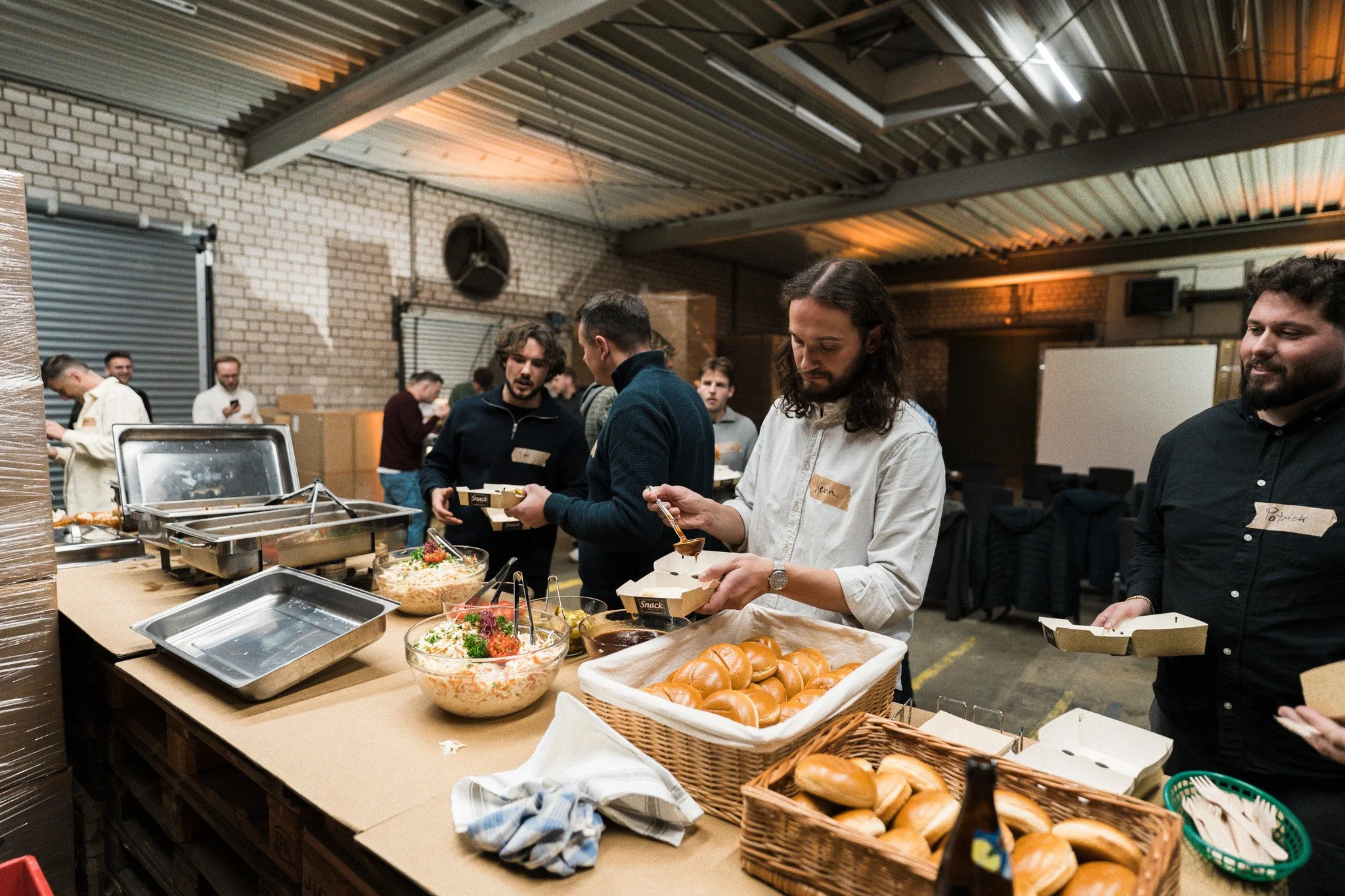 Menschen stehen in einer Halle beim Essen, mit Salat, Brötchen und warmen Speisen auf einem Buffet. Sie bedienen sich und unterhalten sich.