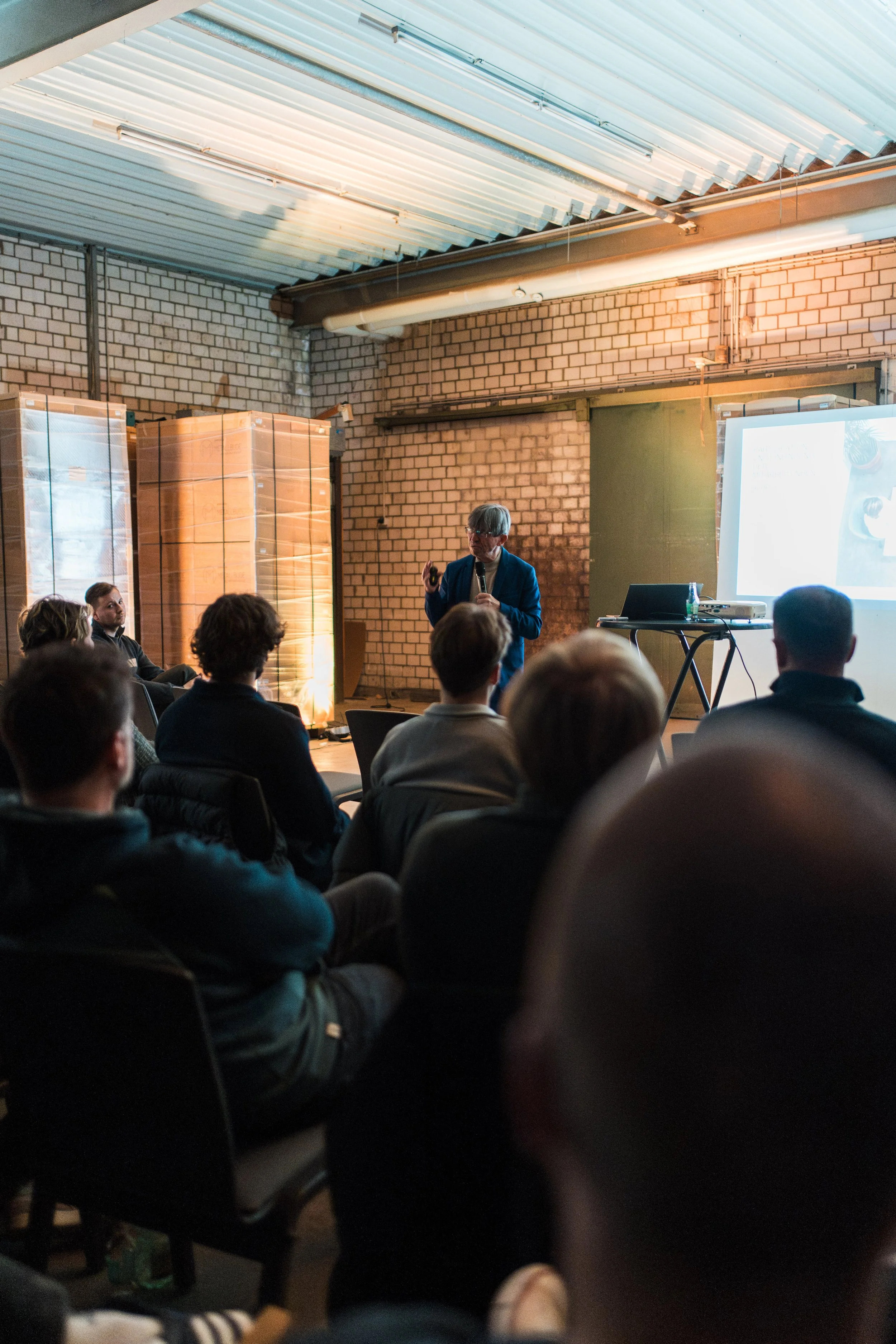 Ein Mann im blauen Anzug hält eine Präsentation vor einem Publikum in einem industriellen Raum mit Ziegelwänden und einer hohen Decke.