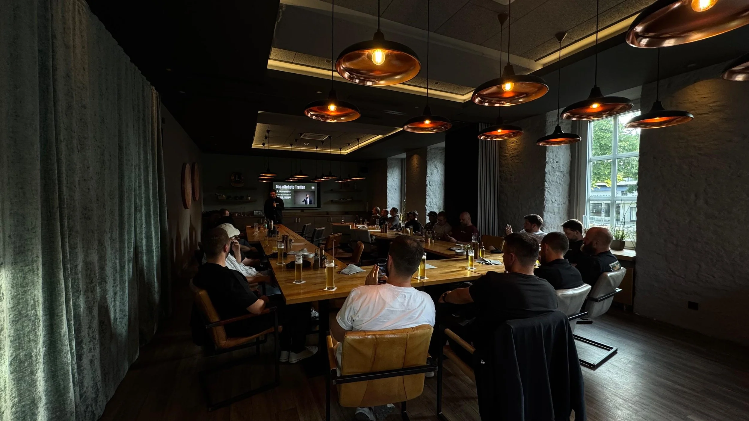 Besprechung oder Präsentation in einem modernen Konferenzraum mit mehreren Teilnehmern, die an einem langen Tisch sitzen und auf einen Bildschirm schauen, während draußen Sonnenlicht durch große Fenster scheint.