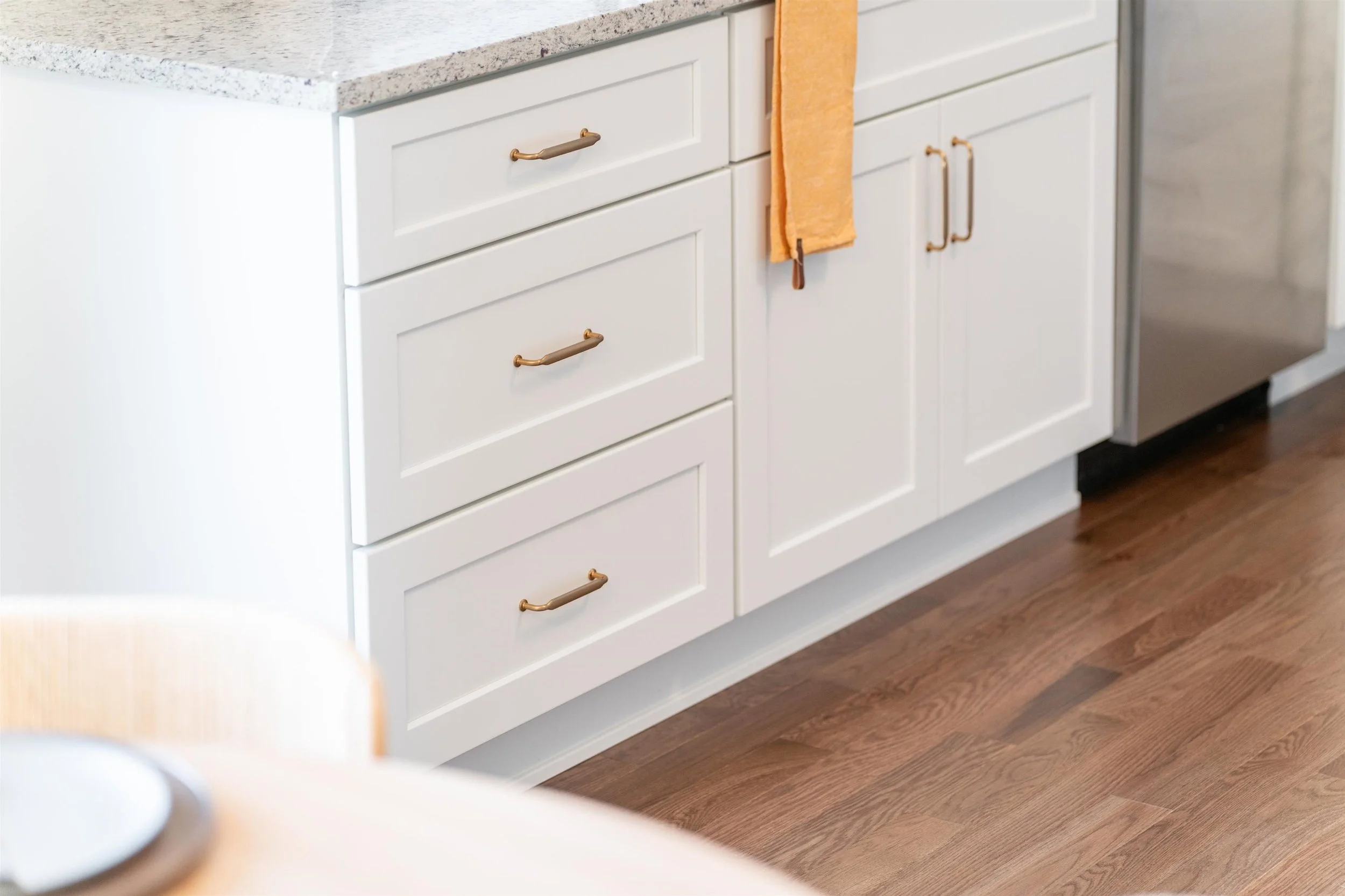 Close-up of white kitchen cabinets with gold handles and a speckled granite countertop, with a towel hanging from one of the cabinet handles.