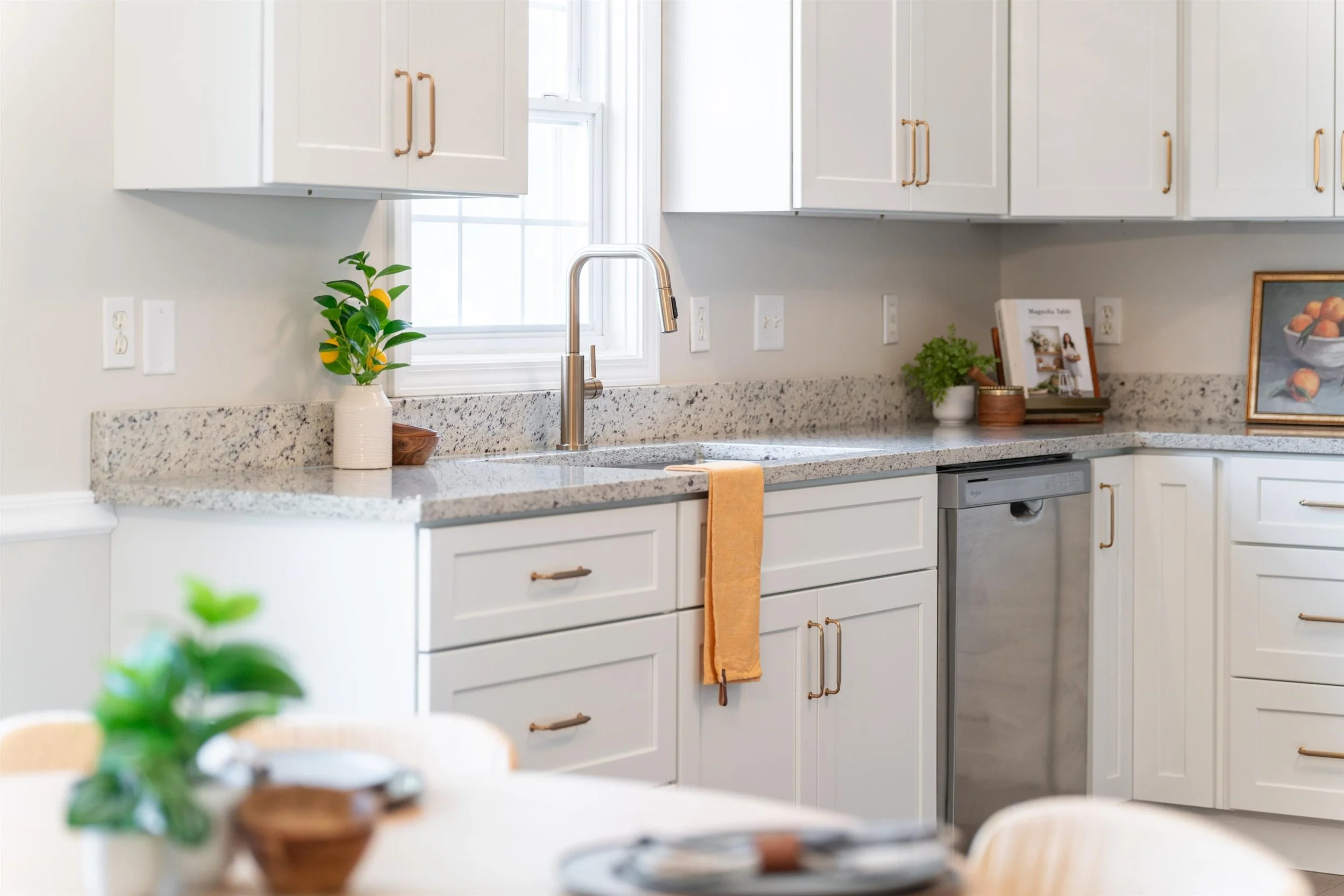 Modern kitchen with white cabinets, granite countertops, a stainless steel dishwasher, a chrome faucet, and decorative items including plants and framed pictures.